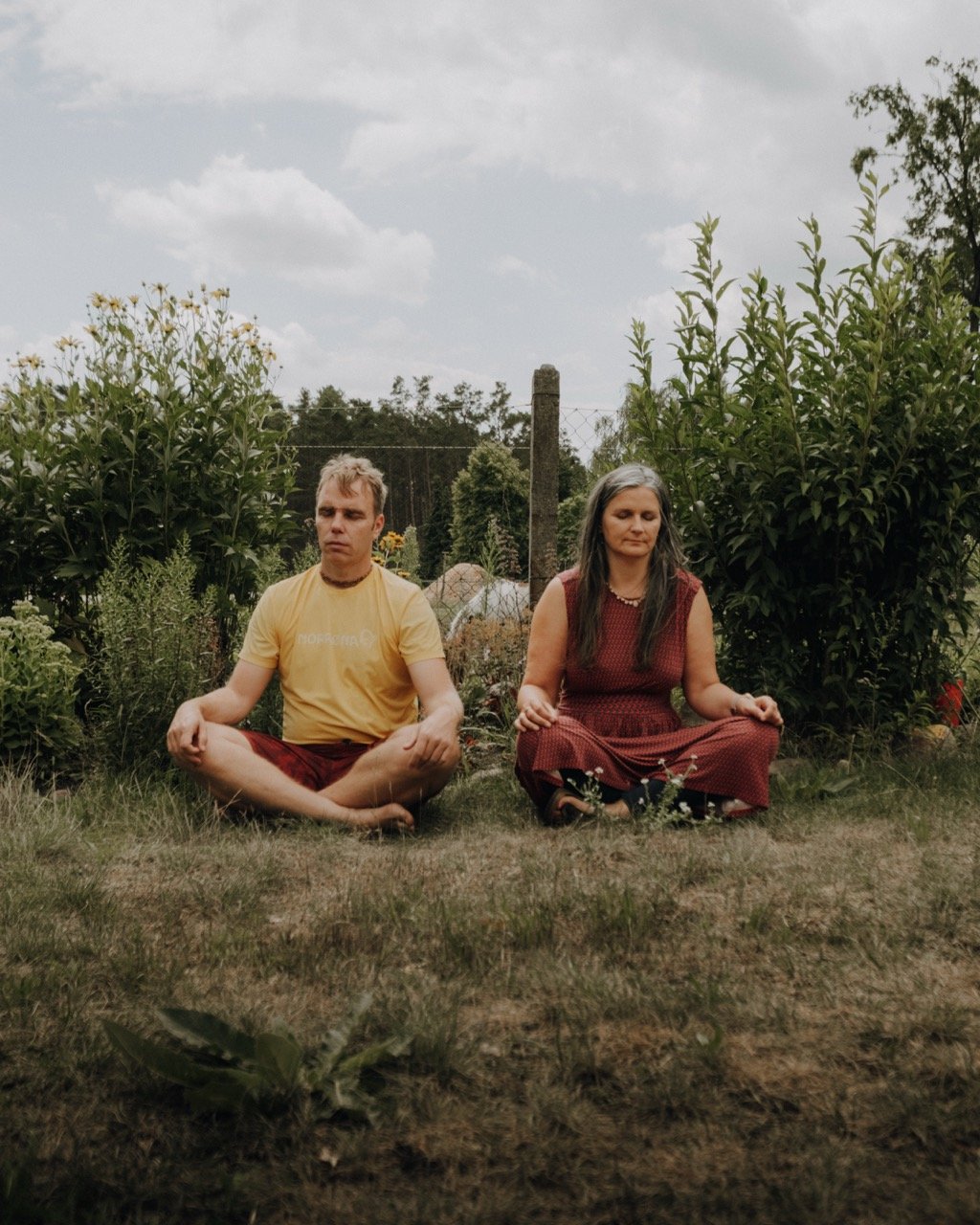 A man and a woman sitting cross-legged on the grass in a garden, with their eyes closed and a peaceful expression, surrounded by lush green bushes and trees.