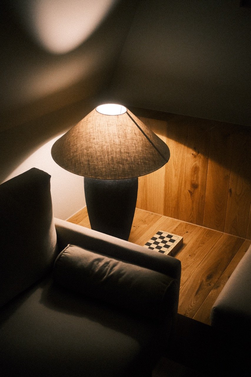 A corner of a room with a lamp on a wooden side table, a sofa with a cushion, and a chessboard on the floor.