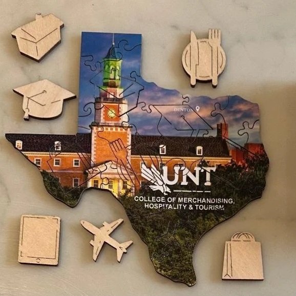 A 25-piece wooden puzzle, with six custom pieces, of the University of North Texas campus in Texas, assembled into a shape of the state, with a box showing the completed puzzle image and the university name and logo - a university gift.