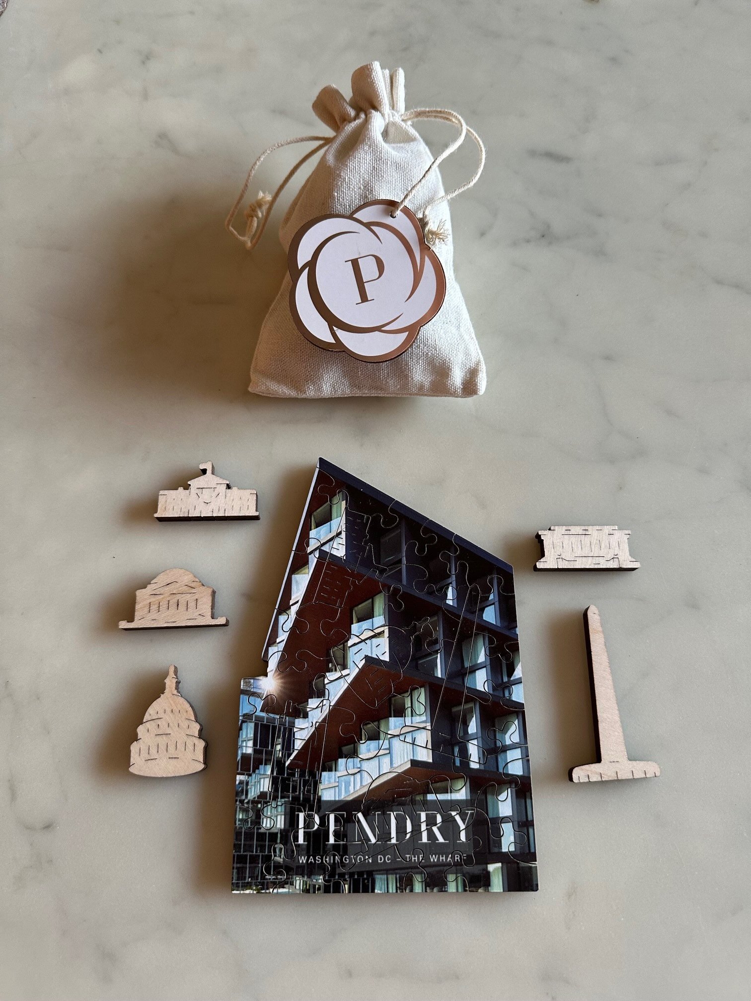 A small beige drawstring bag with a 'P' emblem hangs on a string, next to a completed mini puzzle of a building labeled 'POUNDY' with the words 'WASHINGTON DC - THE WHARF'. Surrounding the puzzle are four small wooden pieces, each depicting a different building or monument.