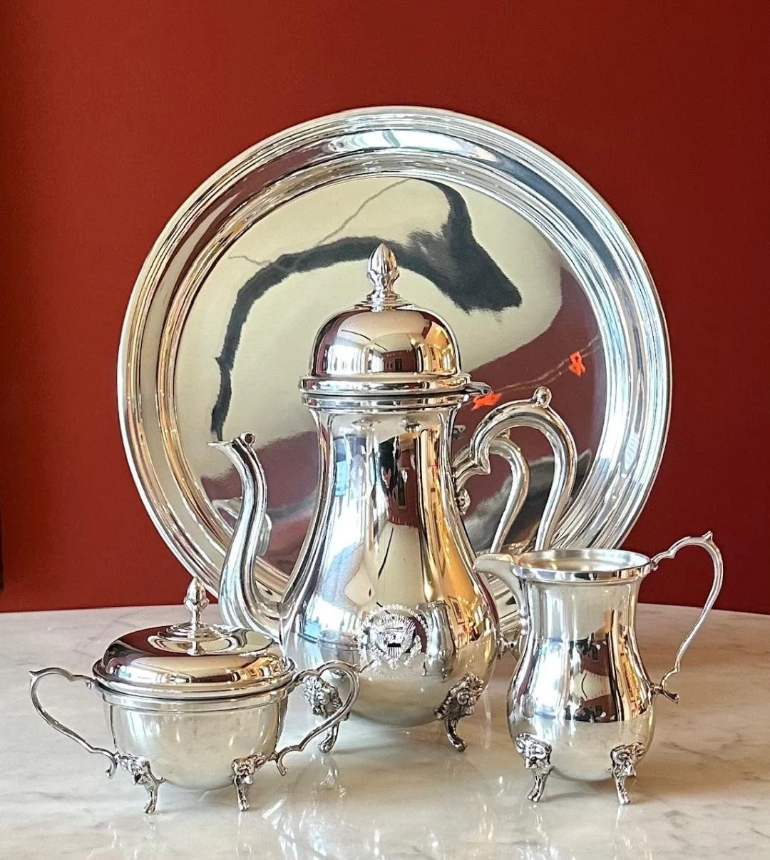 A set of silver tea or coffee serving pieces, including a teapot with a round lid and ornate handle, a sugar bowl with two handles and a matching lid, and a creamer with a curved handle, all positioned in front of a large decorative mirror with a sil