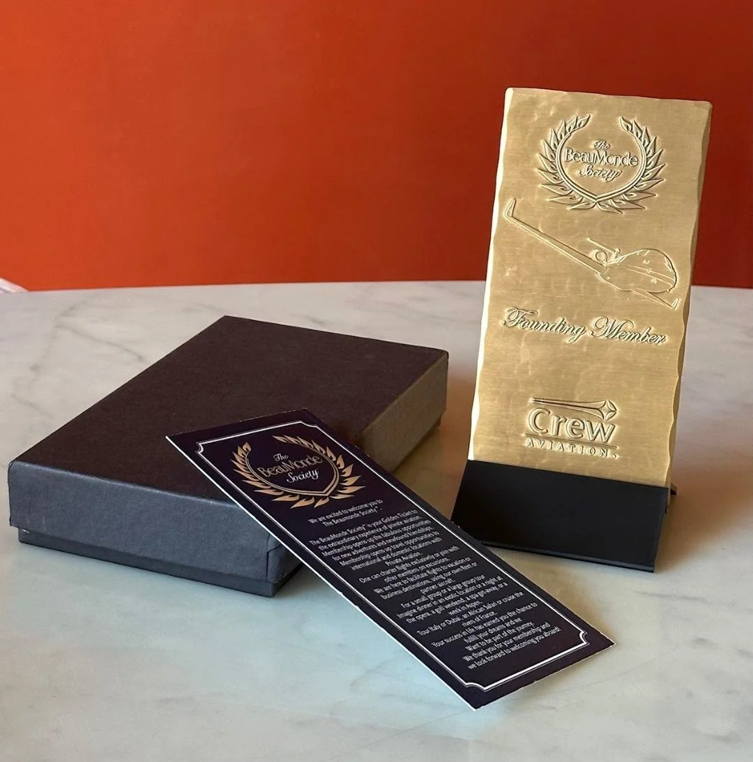 A gold-colored plaque with an embossed golf club and laurel wreath design, reading 'The Beaumonde Society Founding Member' and 'Crew Aviation', stands on a black base. A black and gold booklet and a dark box are placed on a white surface with an orange background.