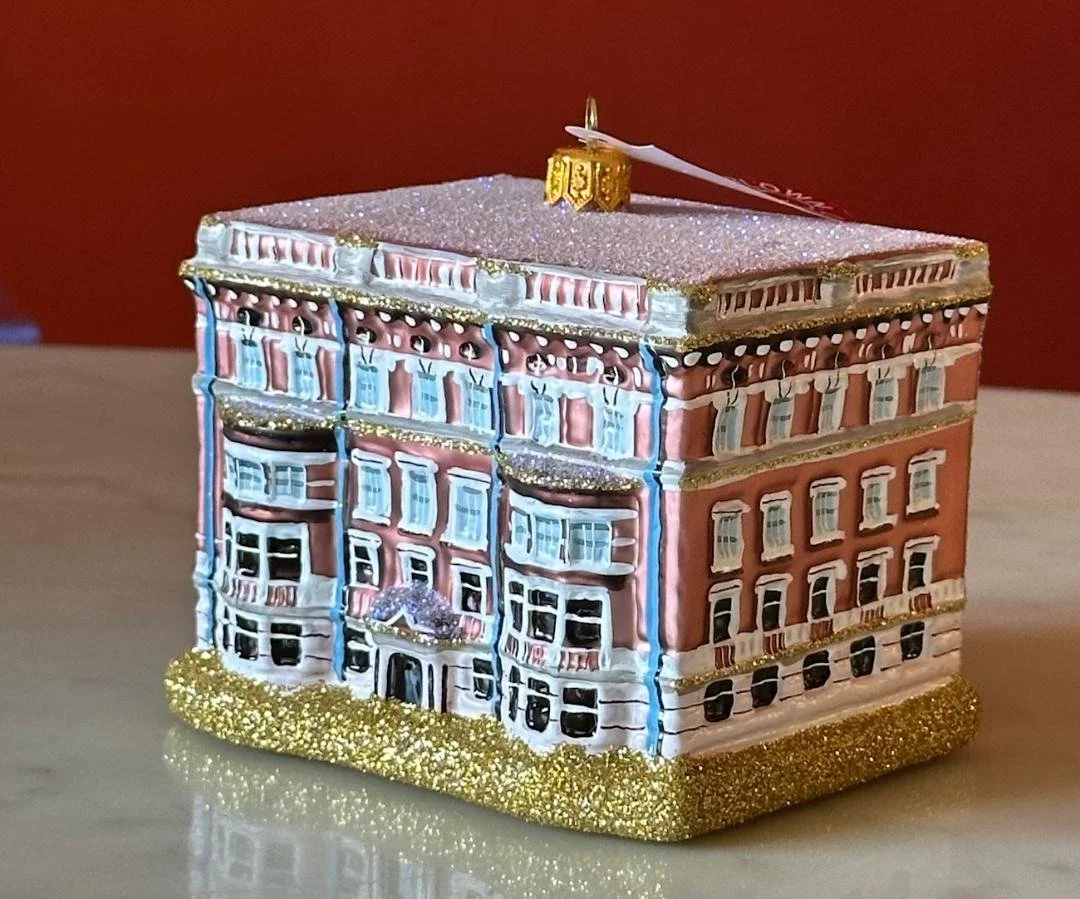 Decorative glass house ornament resembling a historic building (hotel/private club) with detailed architectural features, gold glitter base, and a small tag on top.