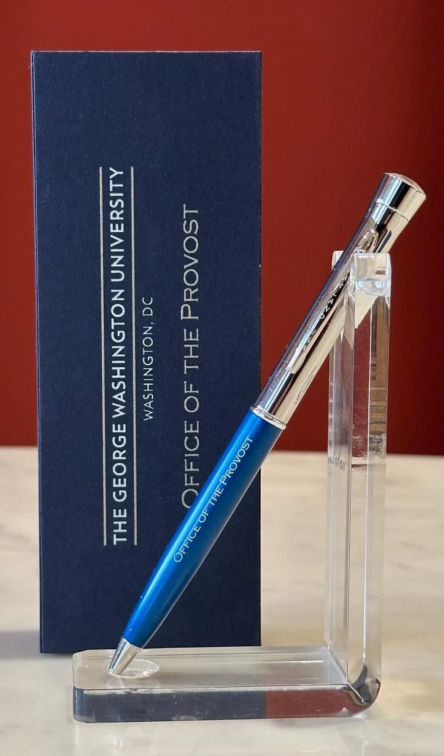 A blue pen with 'Office of the Provost' printed on it, displayed on a clear stand in front of a navy box with the same text and a red background.