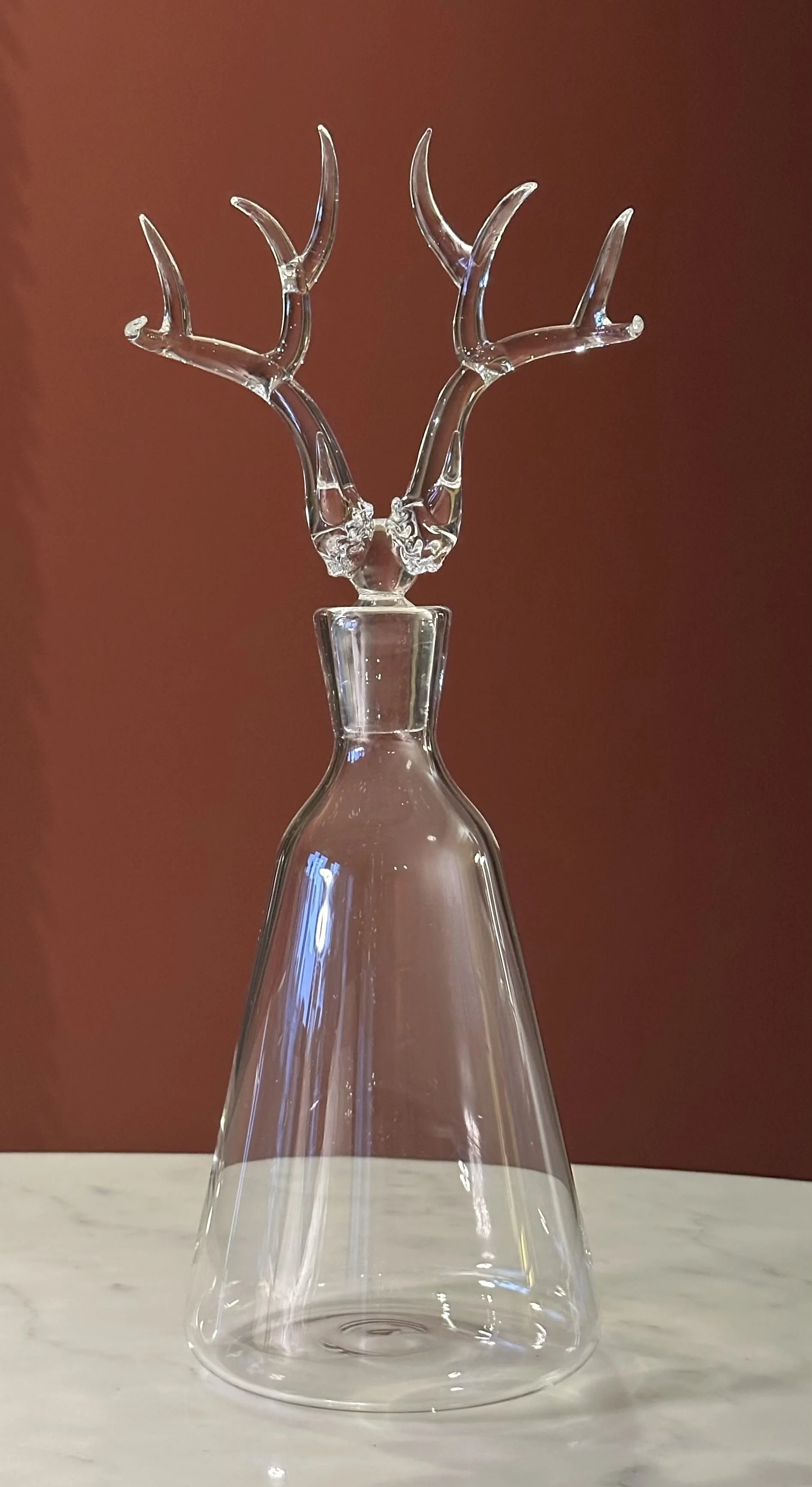 Clear glass vase with decorative glass antlers on top, placed on a white marble surface with a reddish-brown background.
