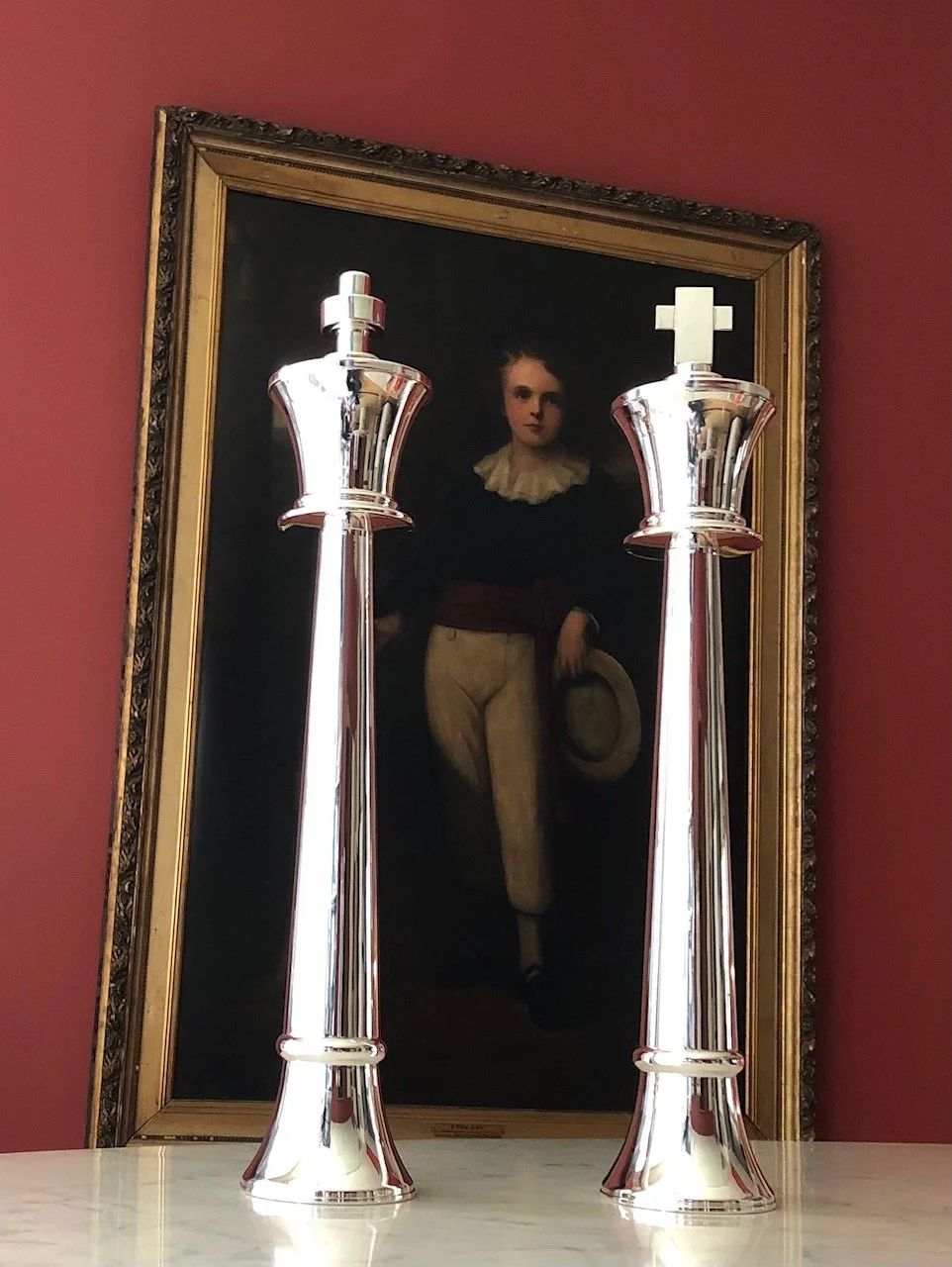 Two tall, shiny silver chess (king and queen) candle holders sit on a white surface in front of a framed portrait of a woman in vintage clothing, holding a hat. The background wall is painted a deep red.