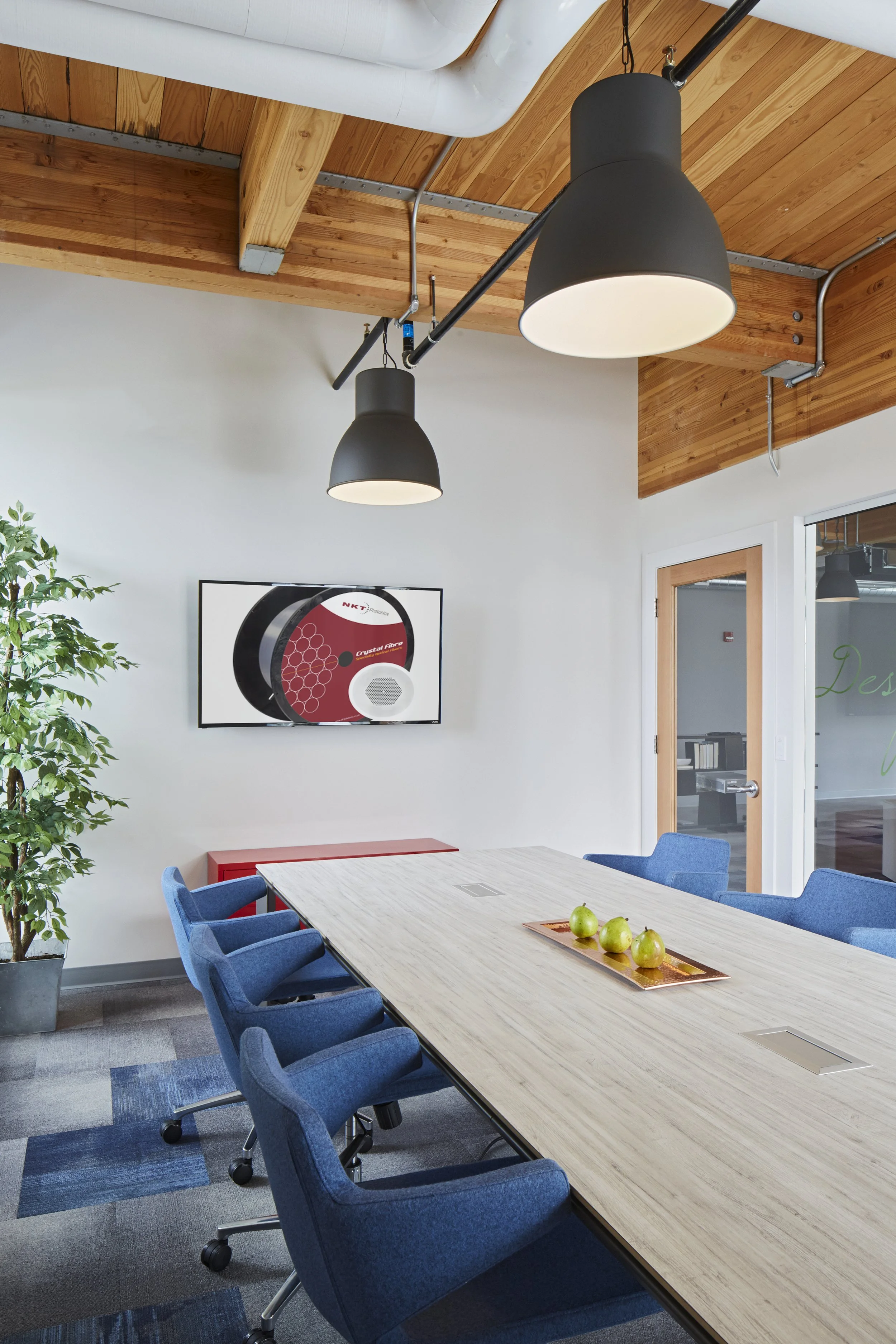 A conference room with a light wooden table surrounded by blue cushioned chairs on a gray and blue carpet. On the table, there is a rectangular tray holding four green apples. In the background, there is a white wall with a mounted flat-screen TV dis