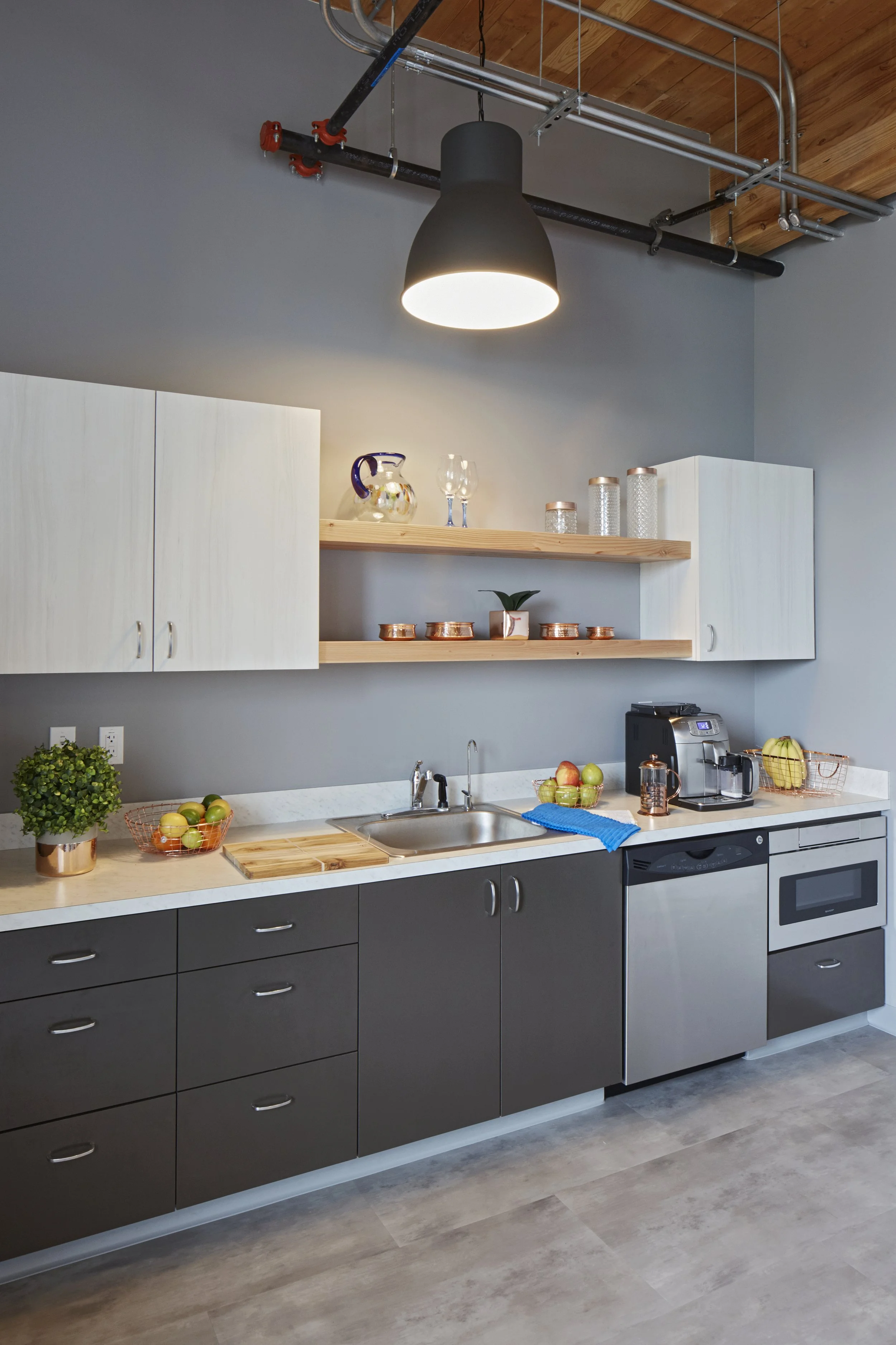 Modern kitchen with gray and white cabinetry, wooden open shelf, black and stainless steel appliances, and decorative items including glass jars, a pitcher, glasses, and bowls of fruit on the countertop, with exposed ceiling pipes and wooden ceiling.