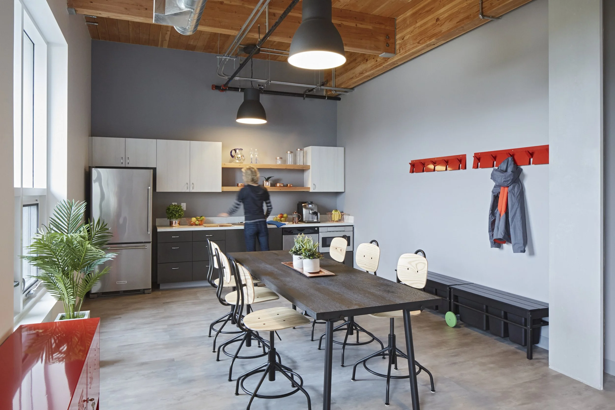 Modern kitchen and dining area with wooden ceiling, gray walls, large table with chairs, kitchen cabinets, plants, and simple decor.