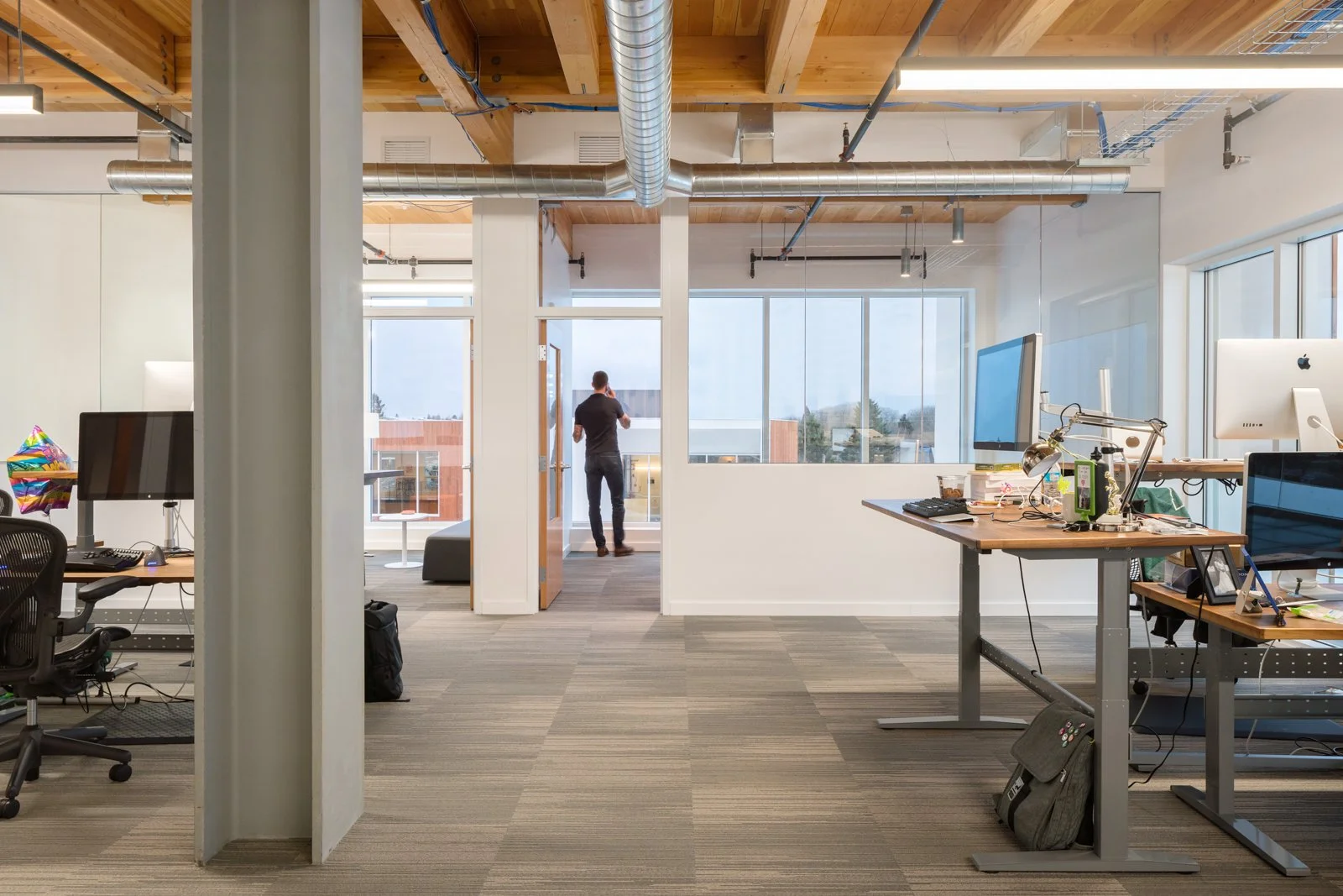 Modern office space with desks, computers, and office chairs. A man stands near glass doors leading to a balcony. The office has exposed wooden ceiling beams, large windows, and industrial ductwork.