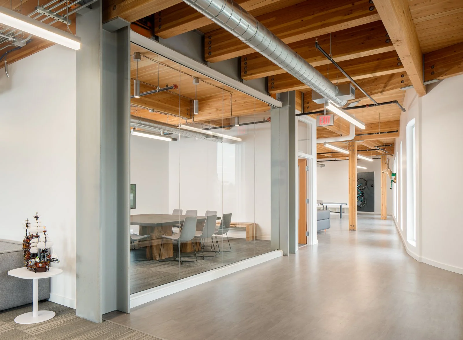 Modern office space with a conference room enclosed by glass walls, featuring a wooden conference table and white chairs, exposed wooden ceiling beams, ductwork, and a hallway with large windows and a few decorative items.