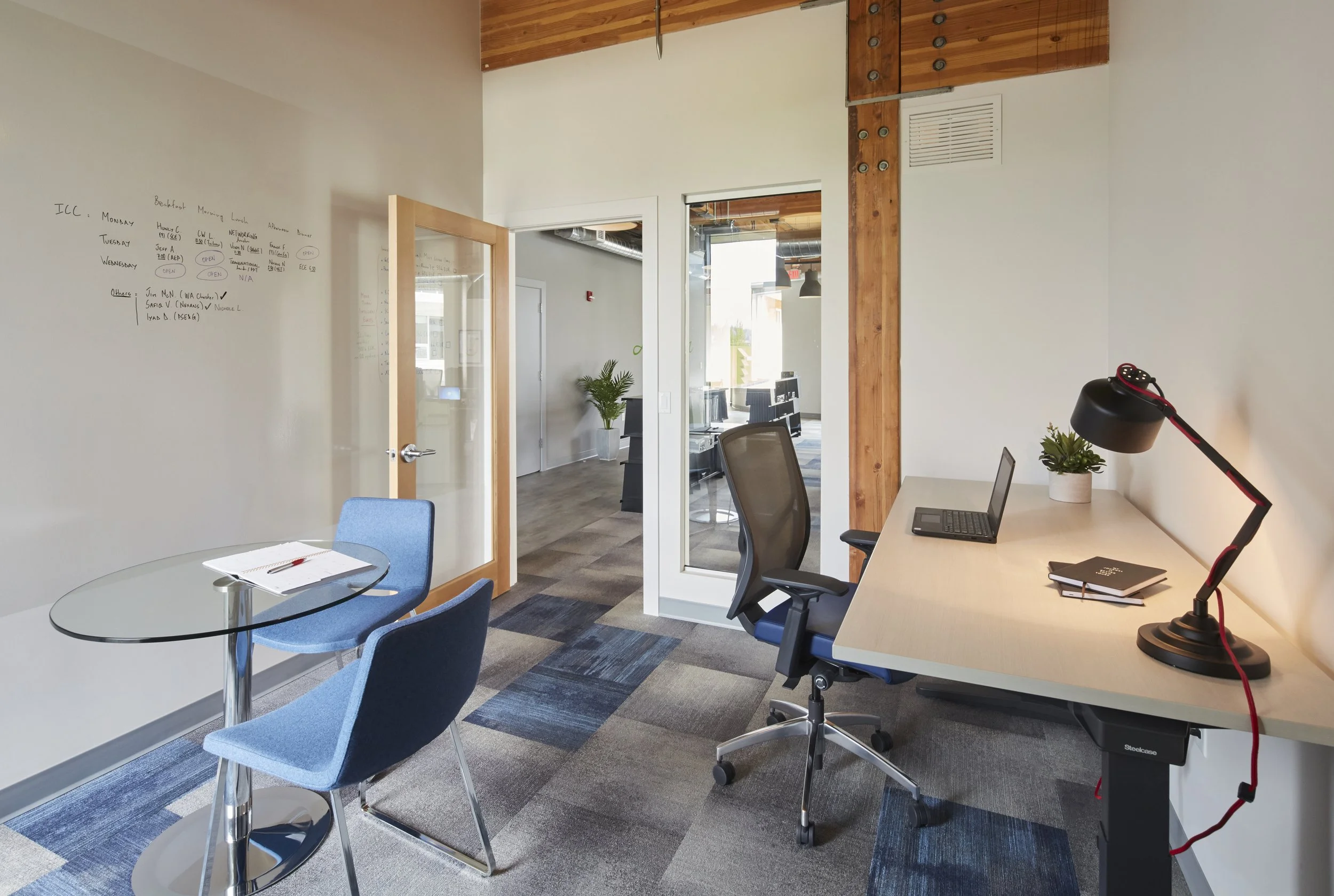 Modern office workspace with a glass meeting table and blue chairs, a white desk with a black office chair, a laptop, a plant, and a black desk lamp. There are glass doors leading to another room with additional desks and office equipment. The office