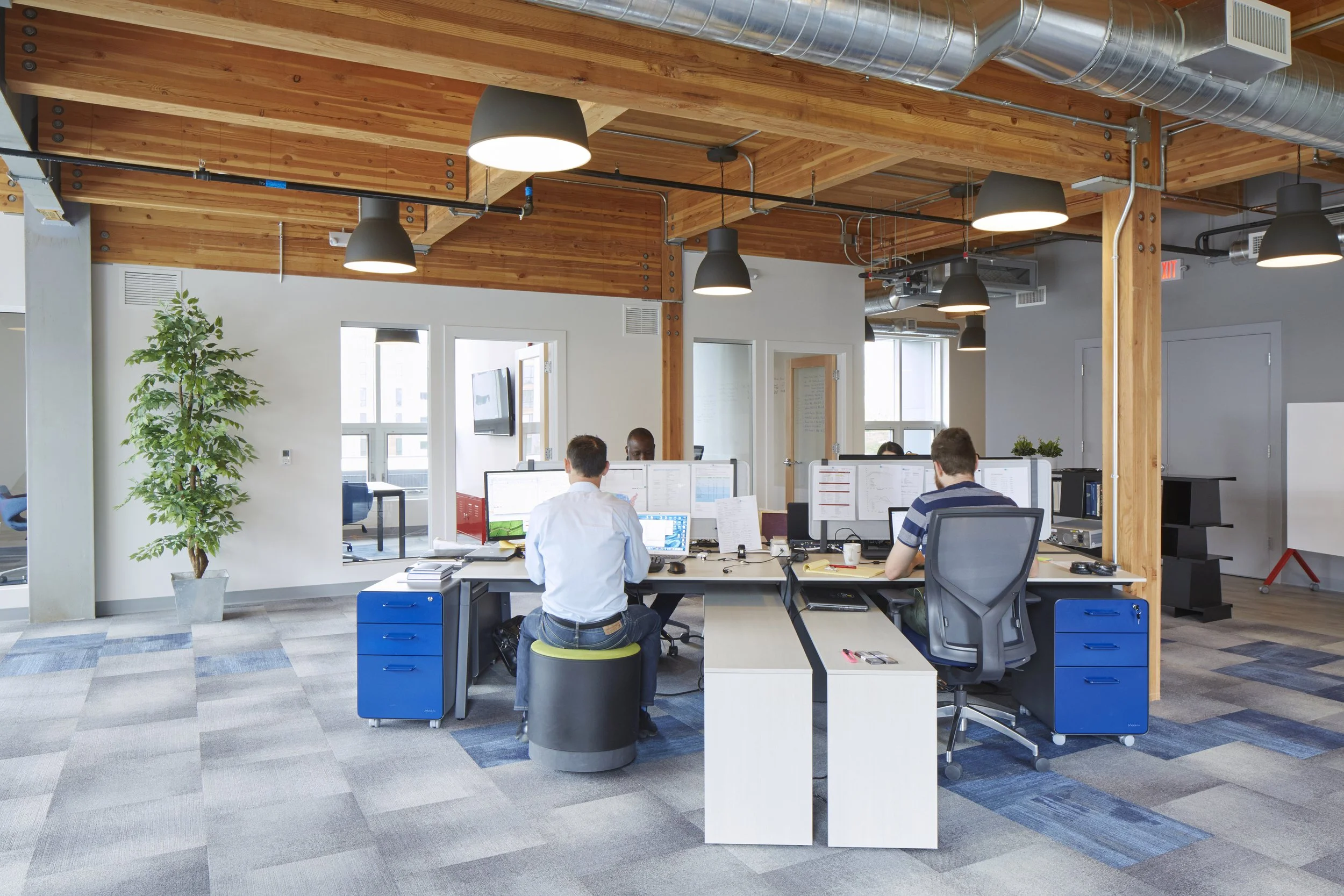 Open-plan modern office with wooden ceiling beams, ductwork, and multiple employees working at desks with computer monitors. Large windows, a potted plant, and adjustable hanging lights are visible.