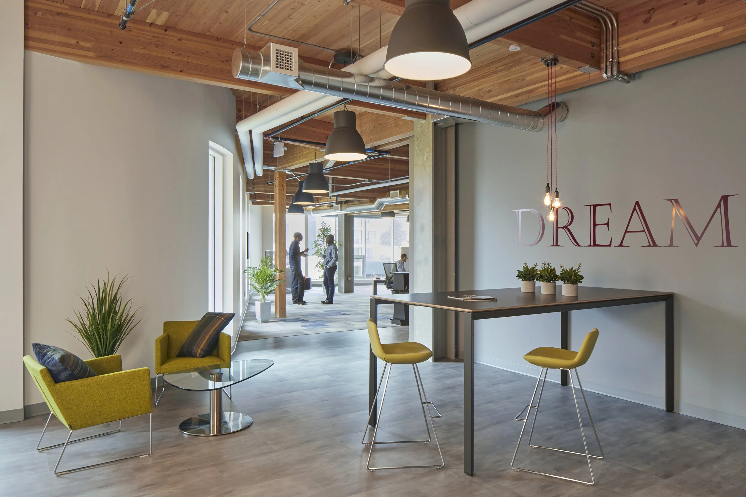 Open office space with modern furniture, ceiling with wooden panels and exposed ducts, two people talking near large windows, plants, and a decorative wall with the word 'DREAM' on it.