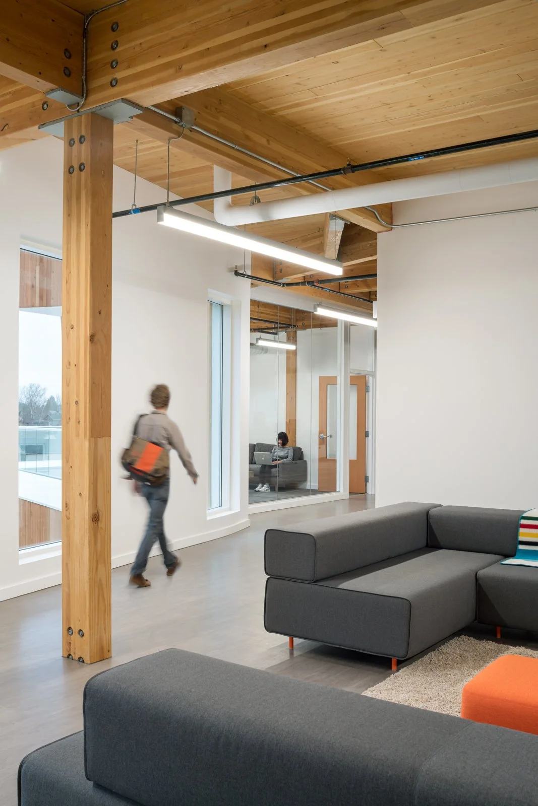 Modern office lounge with wooden ceiling, white walls, and gray sofas, one person walking with a bag and another sitting with a laptop in a glass-enclosed room.