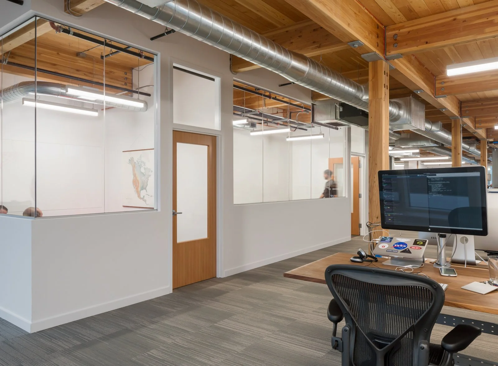 Modern office workspace with a wooden ceiling, exposed ductwork, glass-walled meeting rooms, a desktop computer, and an office chair.