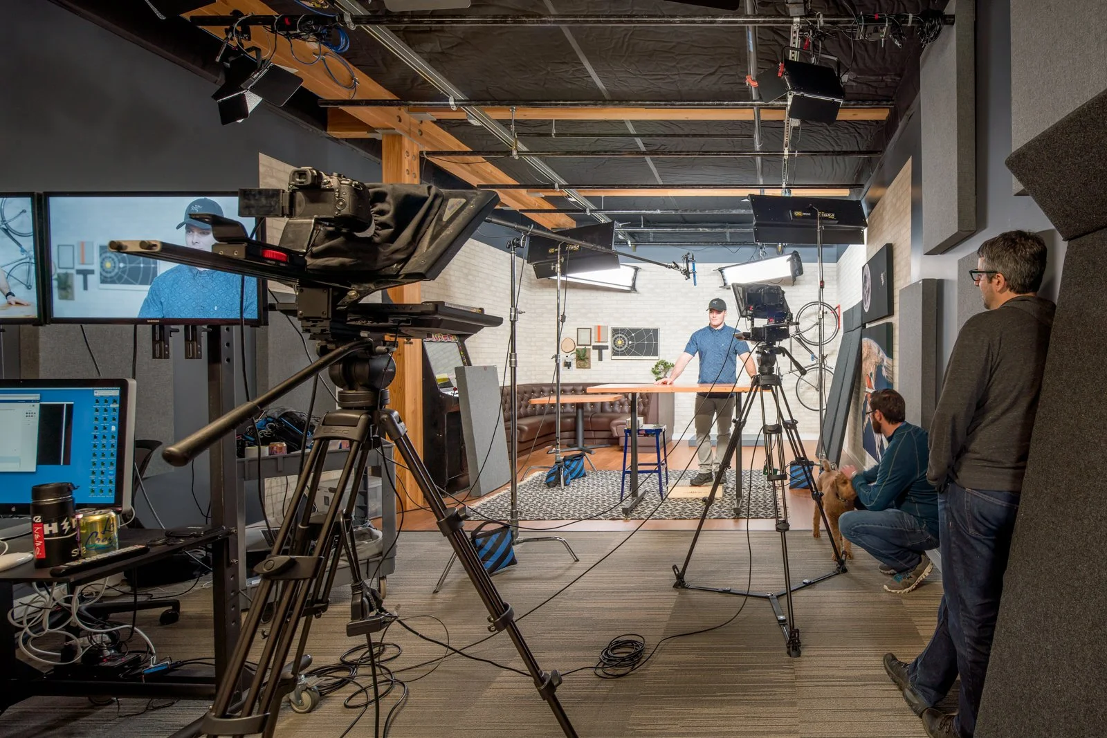A behind-the-scenes view of a video or photo shoot set up in a studio. There is a camera on a tripod, two large softbox lights, and two monitors displaying the subject, a man in a blue shirt wearing a black cap. Two crew members are on the right side