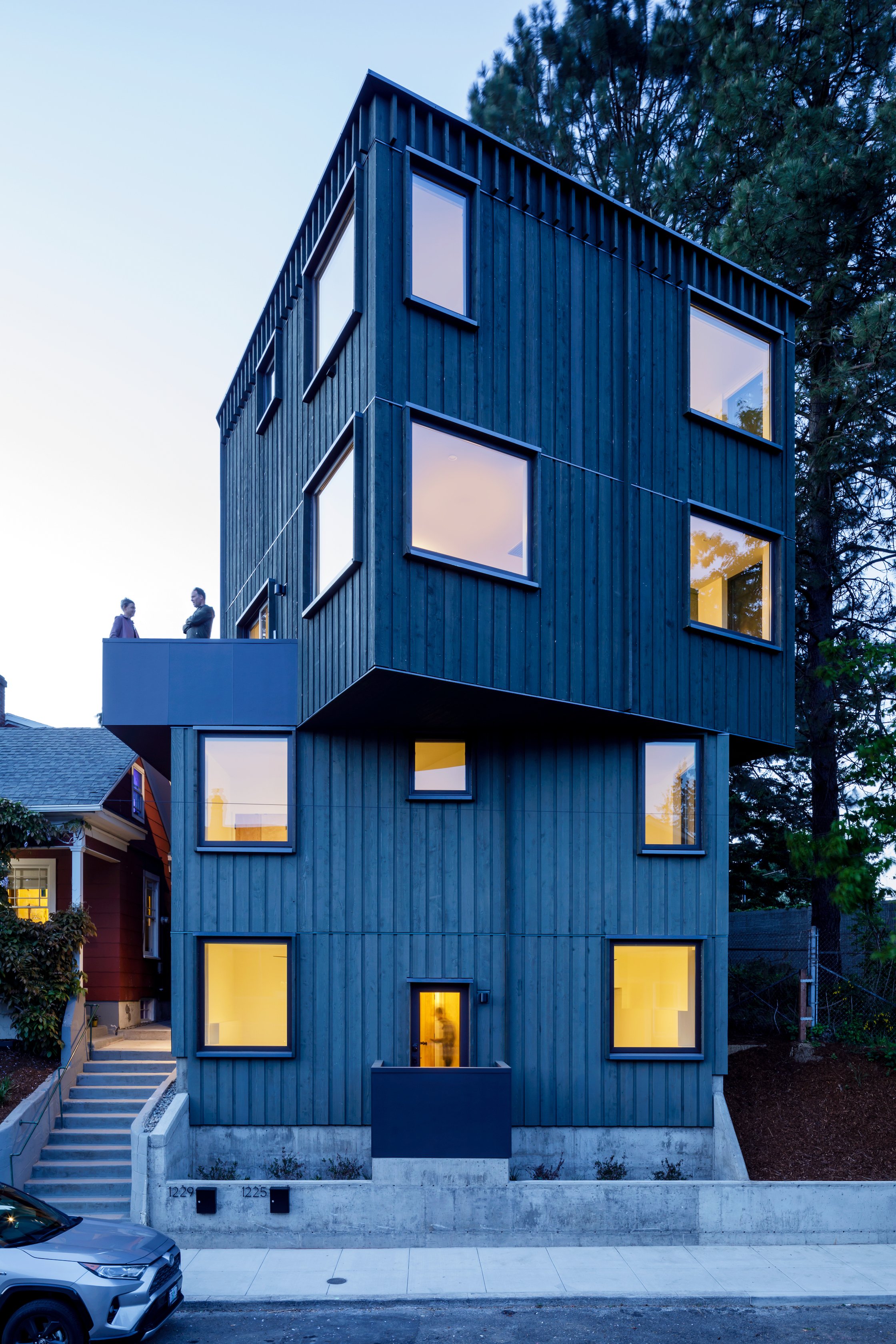 Modern multi-story house with dark wooden exterior and large rectangular windows, illuminated from inside, situated in a residential neighborhood with a car parked outside at dusk.