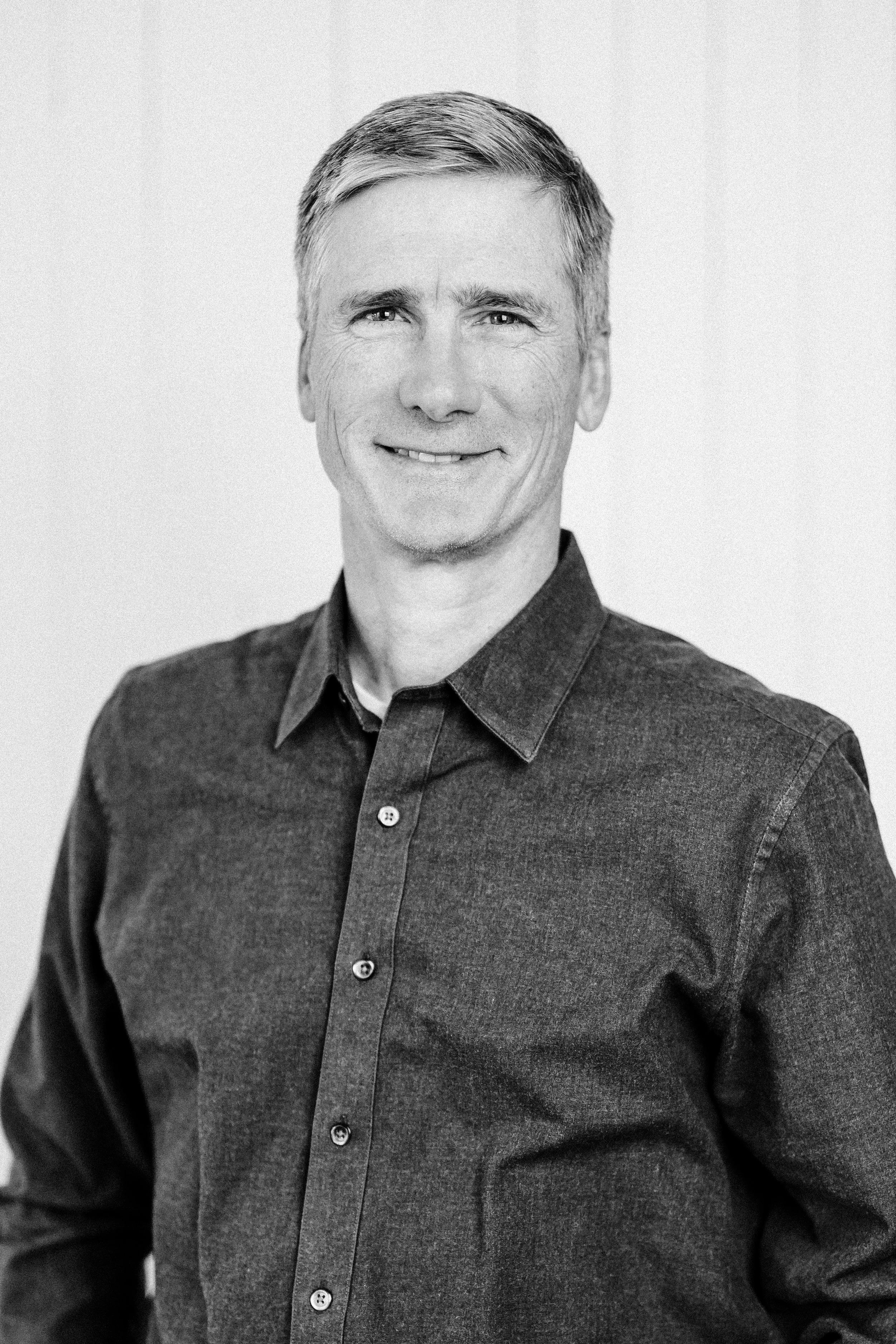 Black and white portrait of a smiling man in a button-up shirt, standing in front of a wooden wall.