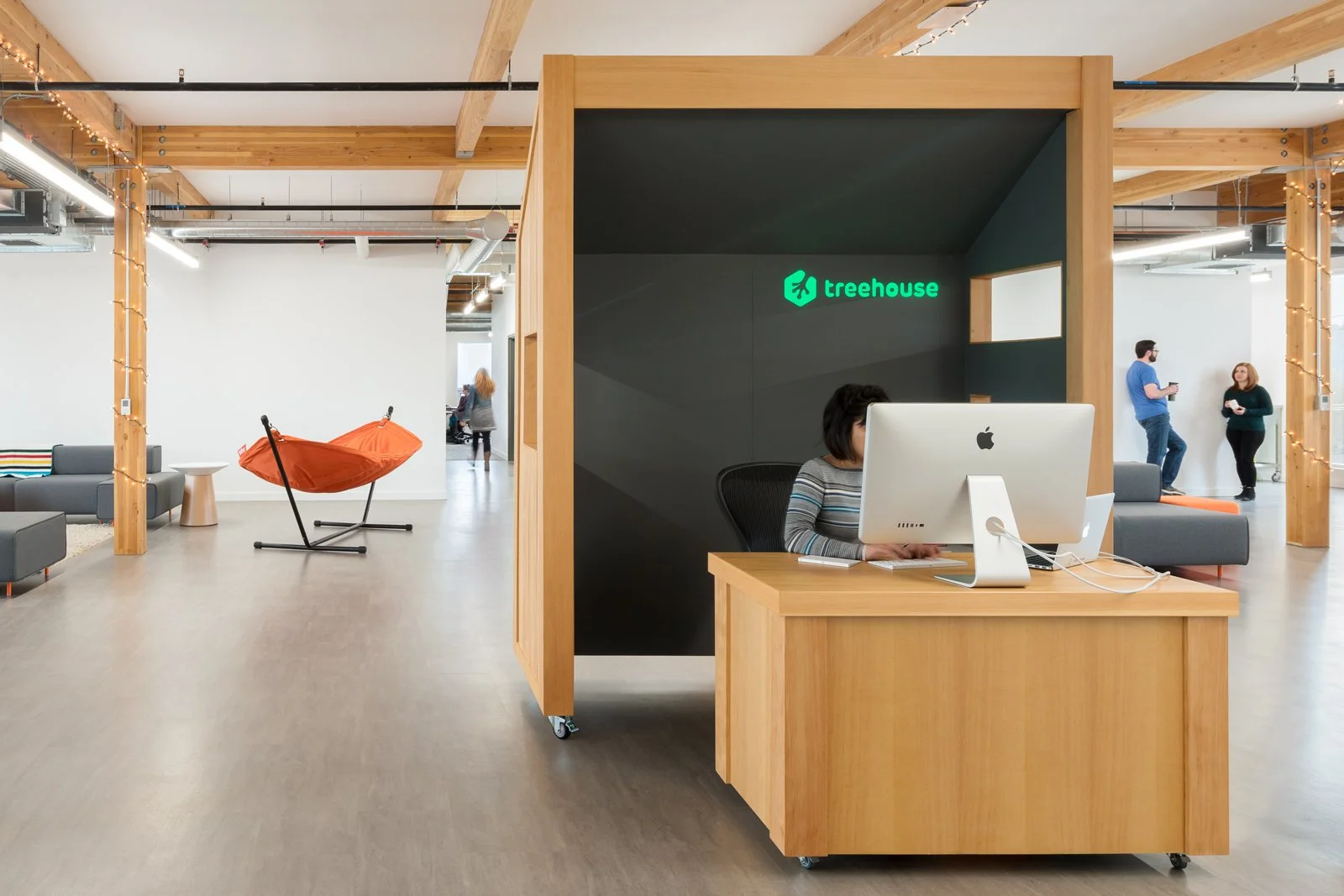 Modern open office space with a reception area, a woman working at a computer inside a wooden booth with a green 'treehouse' sign, seating areas, a hammock chair, and people conversing.