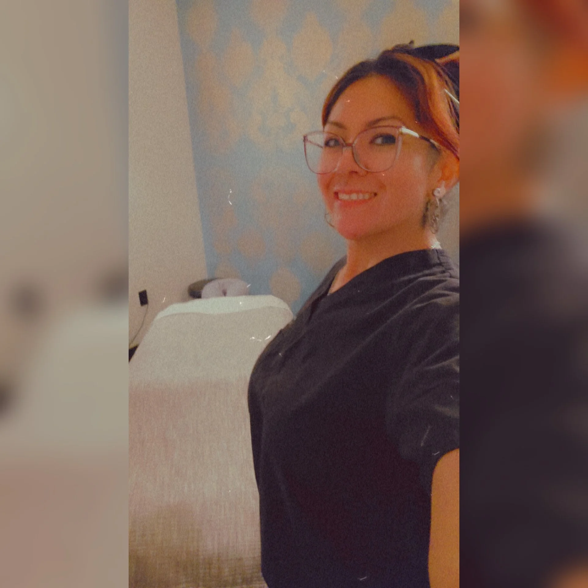 A woman with short hair, wearing glasses, earrings, and a black top, smiling for a selfie in a room with a patterned wall and a piece of furniture in the background.