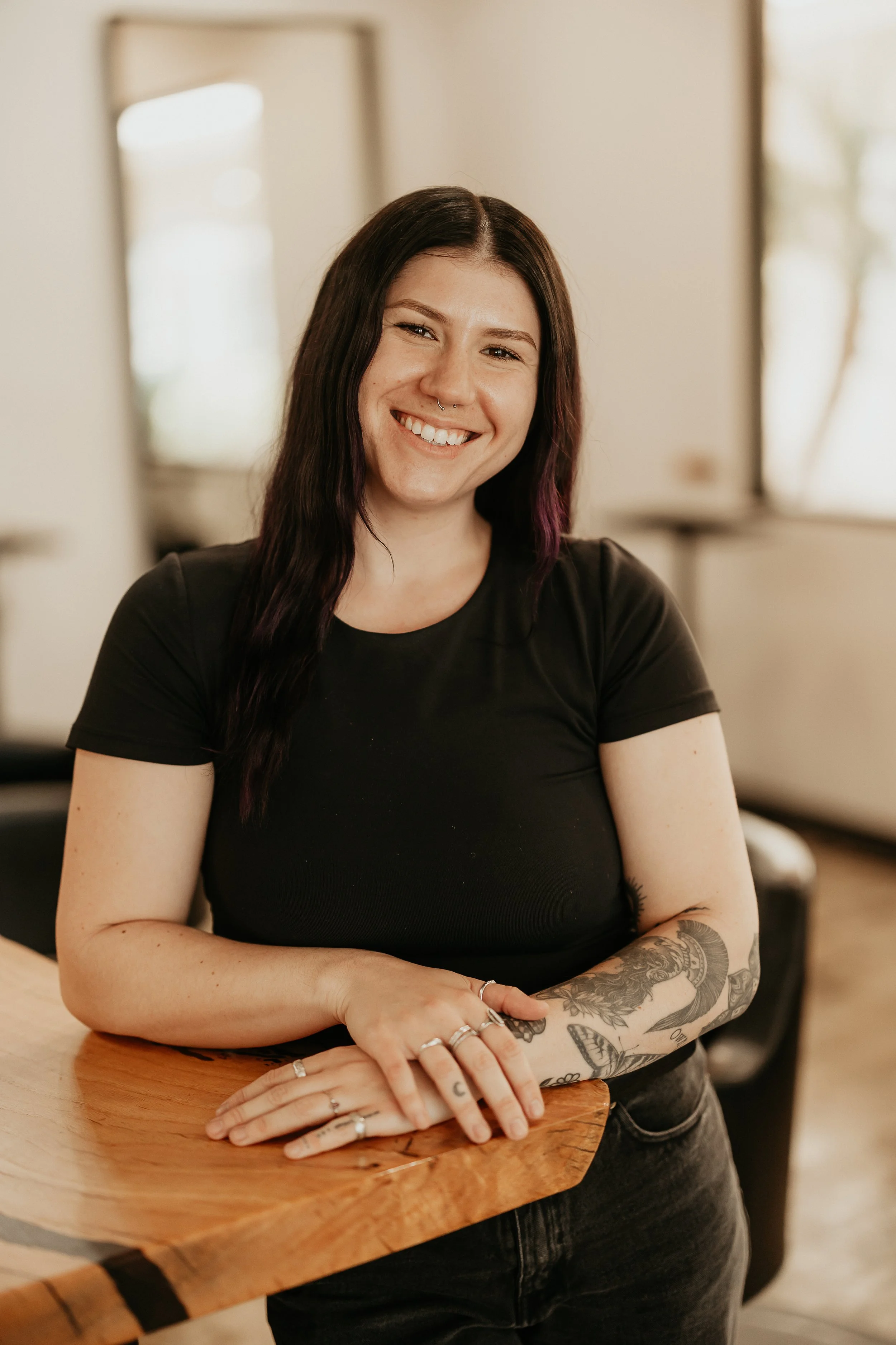 A woman with long dark hair and tattoos on her arm smiling and leaning on a wooden table in a well-lit room.