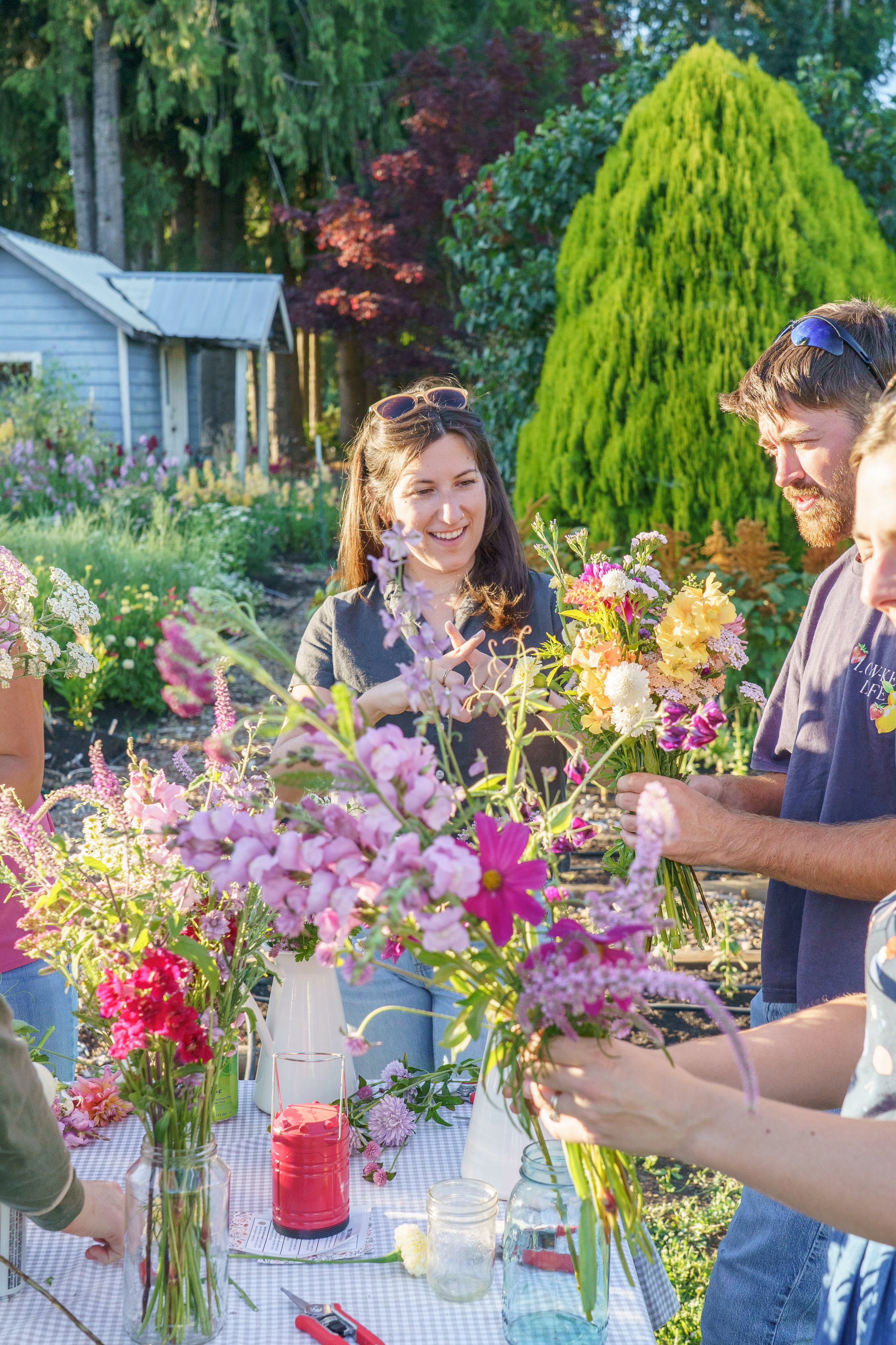 People arranging colorful flower bouquets outdoors in a garden setting with trees and a small shed in the background.