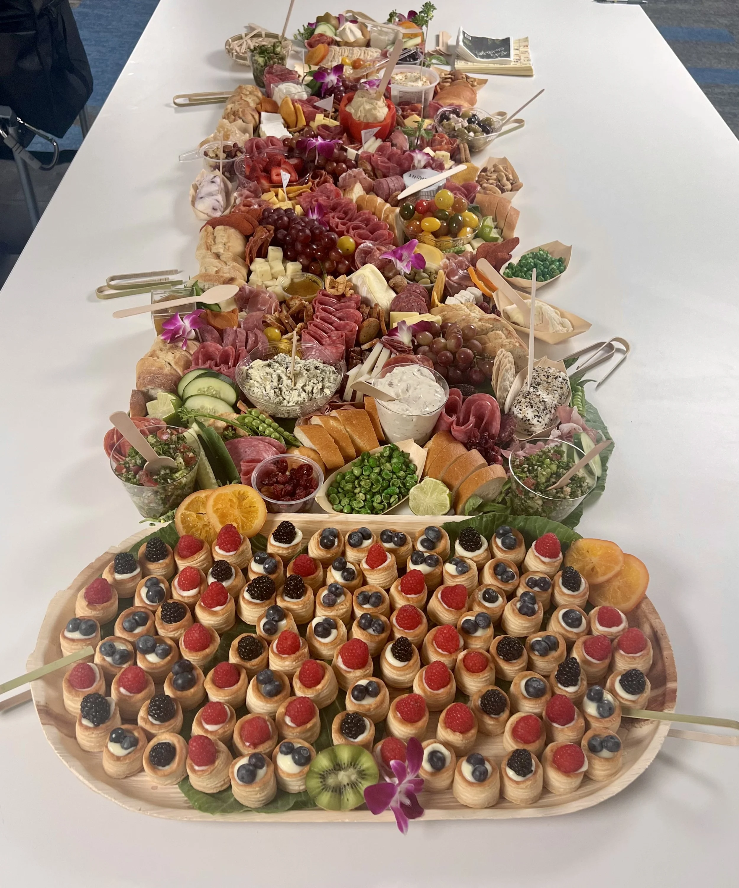 A large platter of assorted mini fruit tarts topped with raspberries, blackberries, and blueberries, arranged on a white table along with an elaborate charcuterie and cheese board featuring meats, cheeses, grapes, olives, crackers, sliced vegetables,