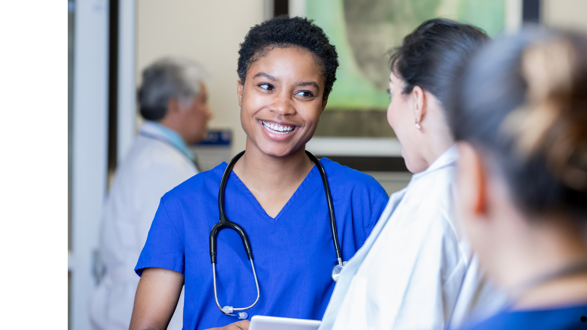 A smiling Black female nurse in blue scrubs with a stethoscope around her neck talking with female colleagues in a medical setting.