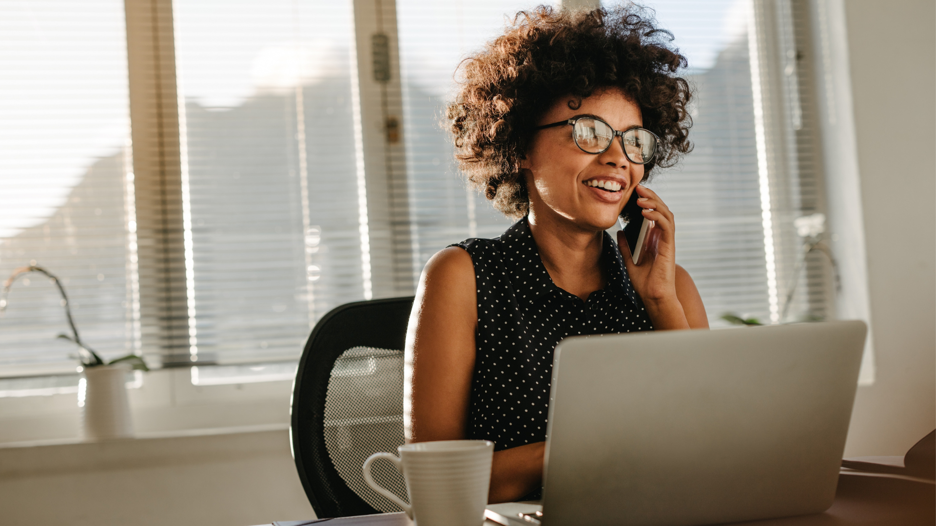 A woman with curly hair and glasses smiling while talking on the phone in an office, sitting in front of a laptop with a cup nearby.