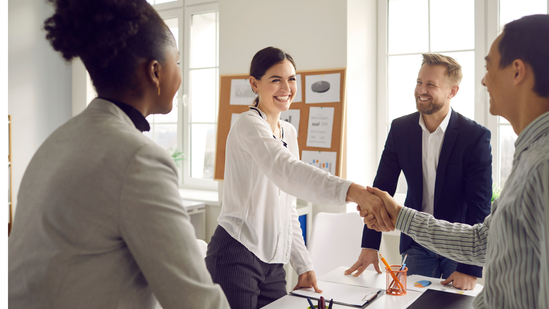 A group of four diverse professionals in an office, with two people shaking hands, smiling, and engaging in a friendly business meeting.
