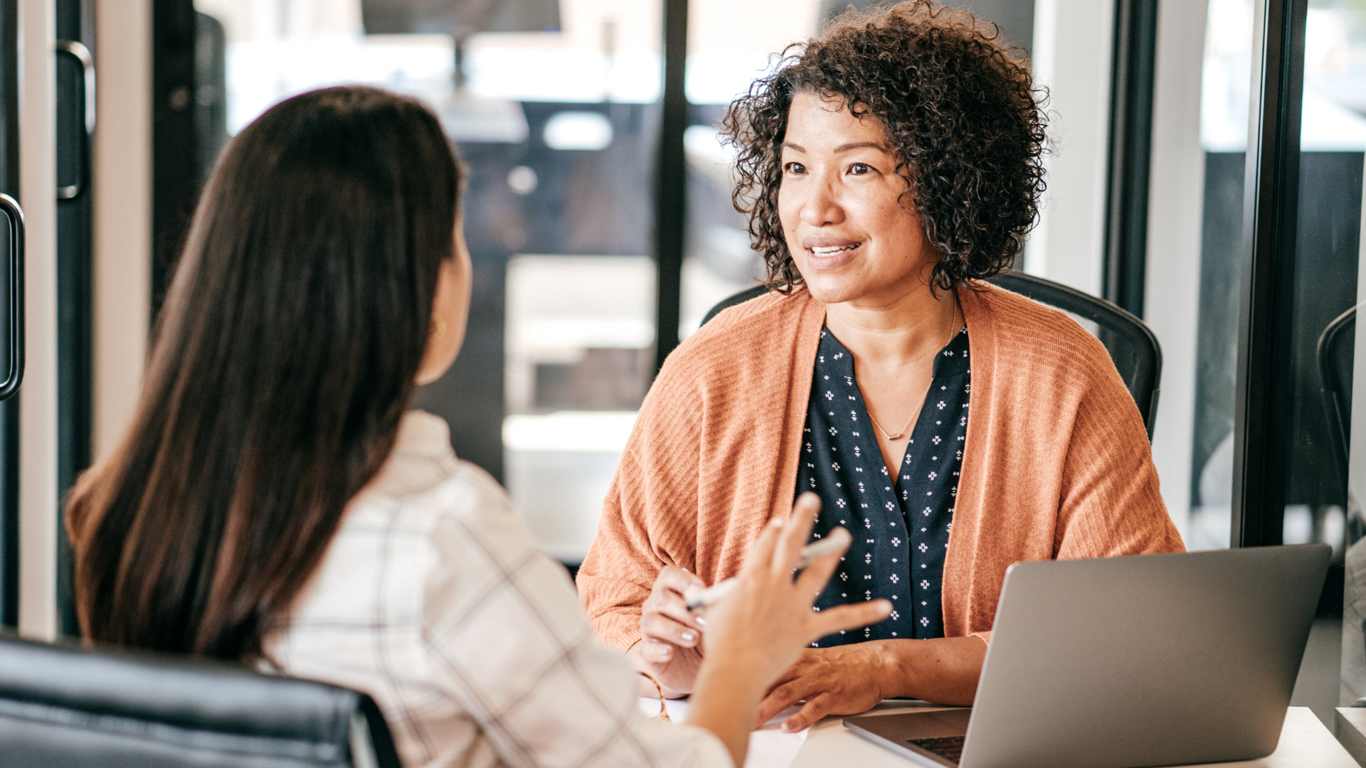 Two women having a discussion in an office setting, with one woman facing away from the camera and the other woman with curly hair facing the camera, sitting at a desk with a laptop.