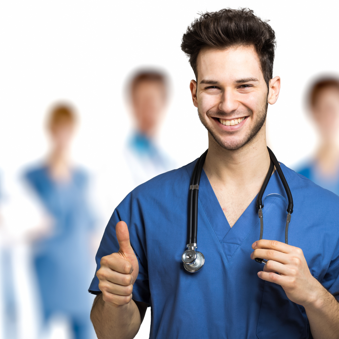 Young male healthcare professional in blue scrubs with stethoscope smiling and giving a thumbs-up, with blurred medical staff in the background.
