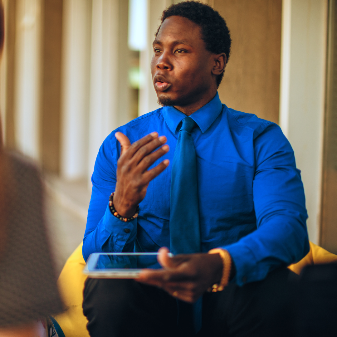 Man in a blue shirt and tie speaking and gesturing with hand while holding a tablet, seated indoors.