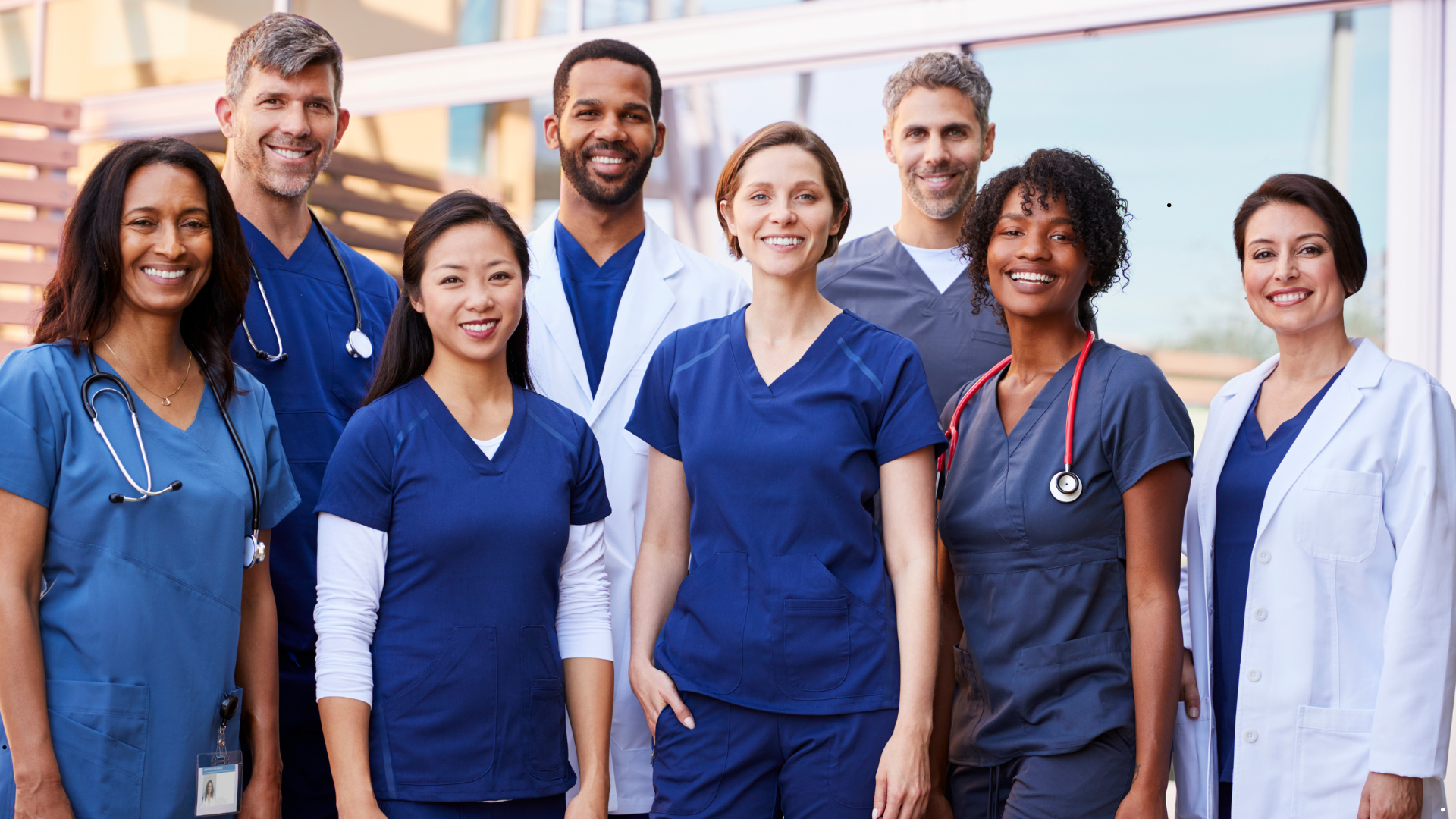 Group of diverse healthcare professionals in scrubs and lab coats standing outdoors smiling at camera.