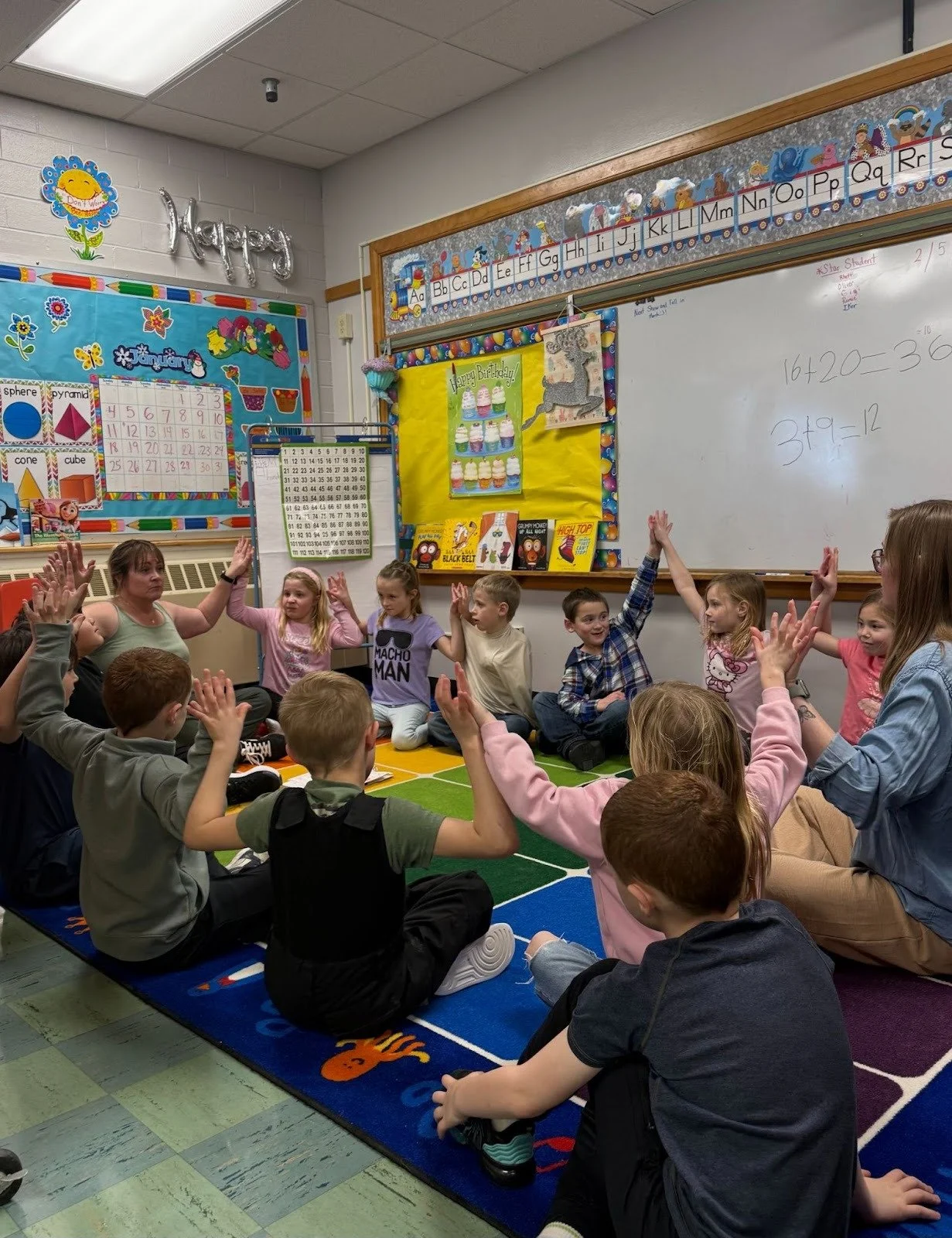A classroom with children sitting on a colorful carpet, raising their hands, and a teacher leading an activity.