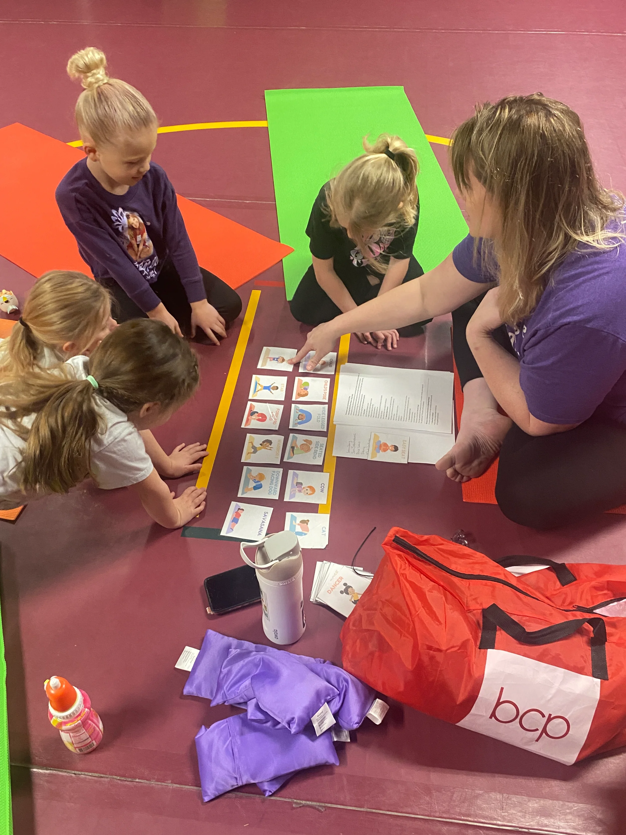 A group of young children and an adult sitting on a gym floor, engaging in an educational activity with cards and printed materials related to kids' yoga. There are colorful yoga mats, a water bottle, a mobile phone, and a large red and white bag nearby.