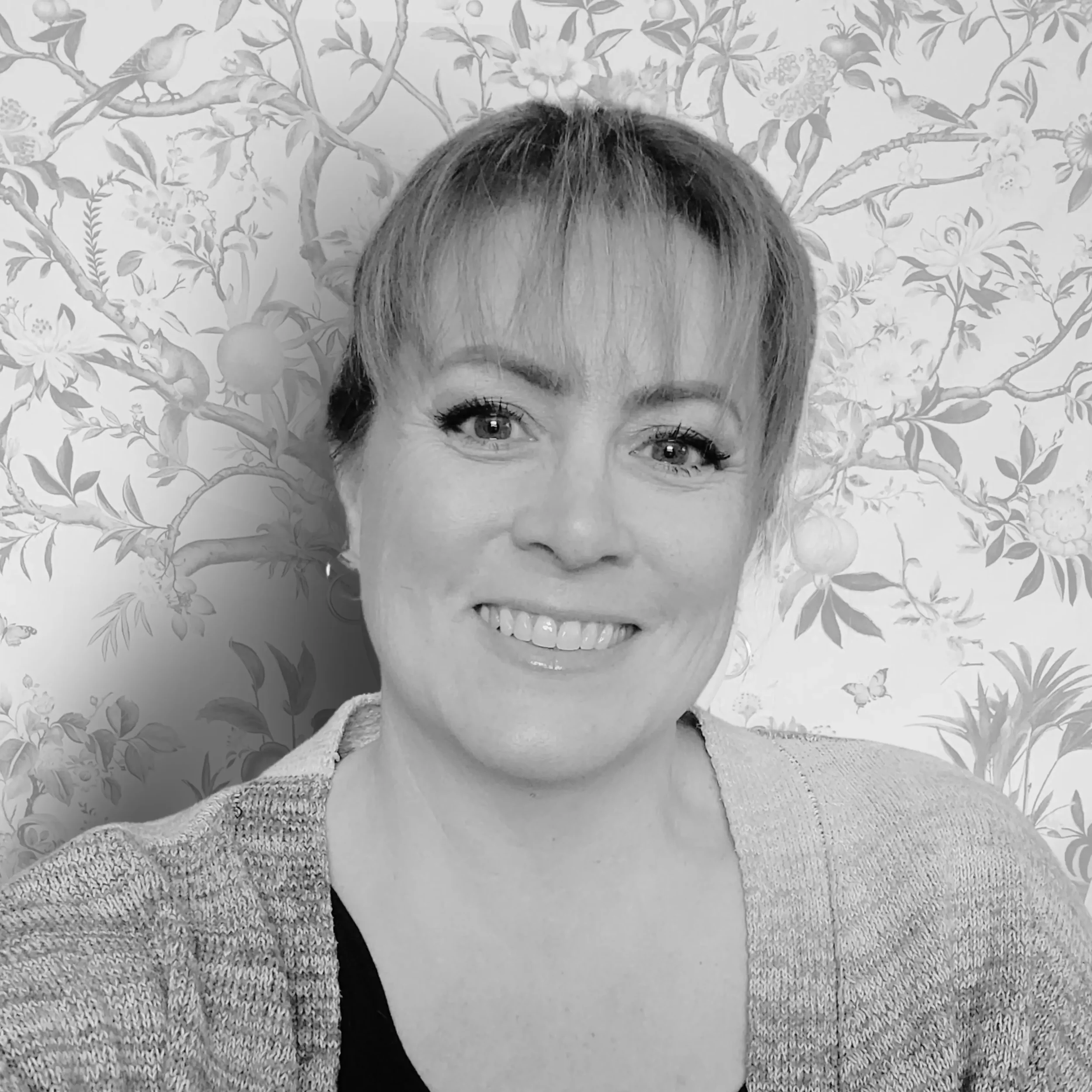 A smiling woman with short hair and wearing a light-colored sweater, standing in front of a floral patterned wallpaper.