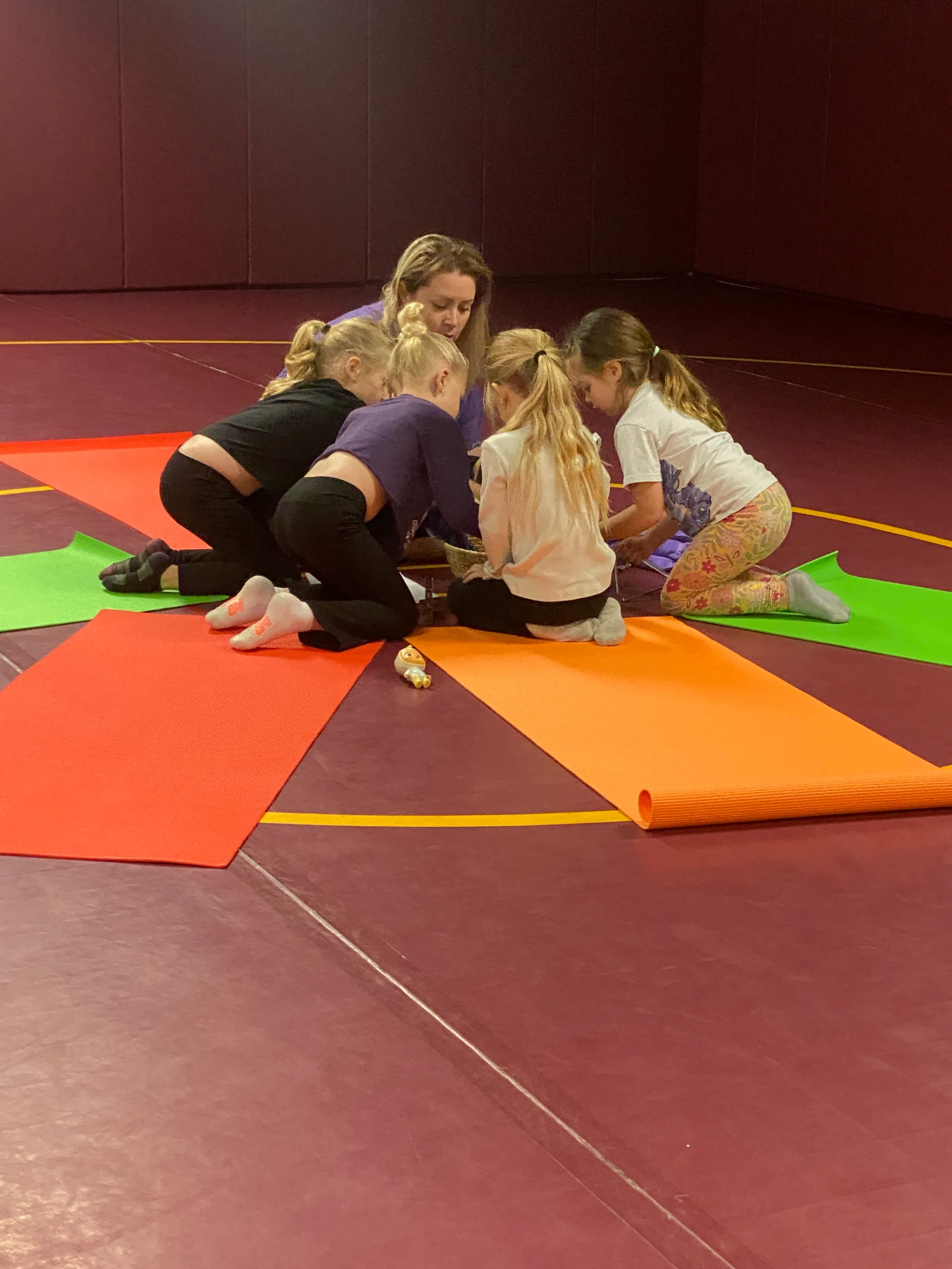 A woman and five young girls sitting on colorful yoga mats on a gym floor, engaged in a group activity or discussion.