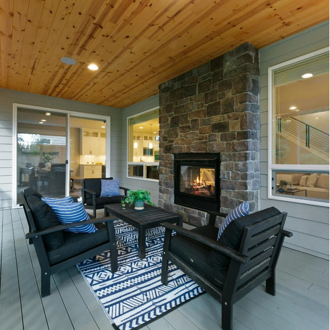 Outdoor patio with wooden ceiling, stone fireplace, and seating area. Modern furniture includes black chairs with blue and white striped pillows, a black table, and a blue patterned rug. Large windows and sliding glass door showcase an interior room.