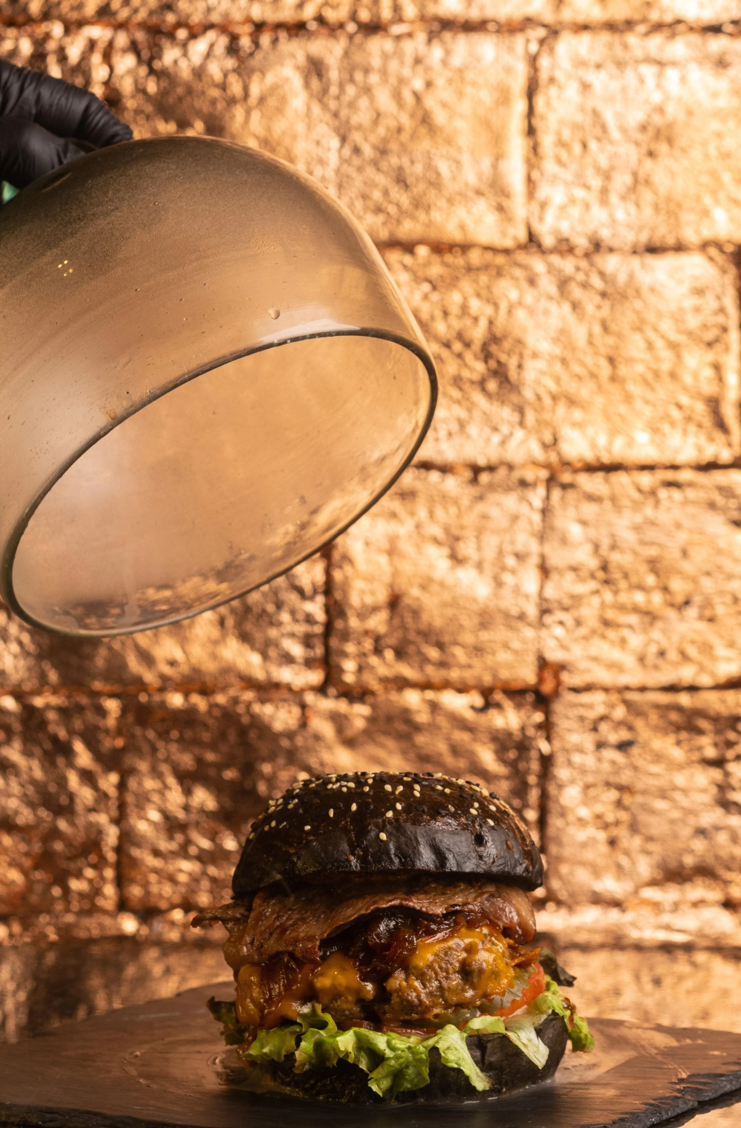 Una hamburguesa con pan negro, carne, vegetales y encima una pieza de queso, sobre una superficie negra y fondo de pared de ladrillos de color marrón.