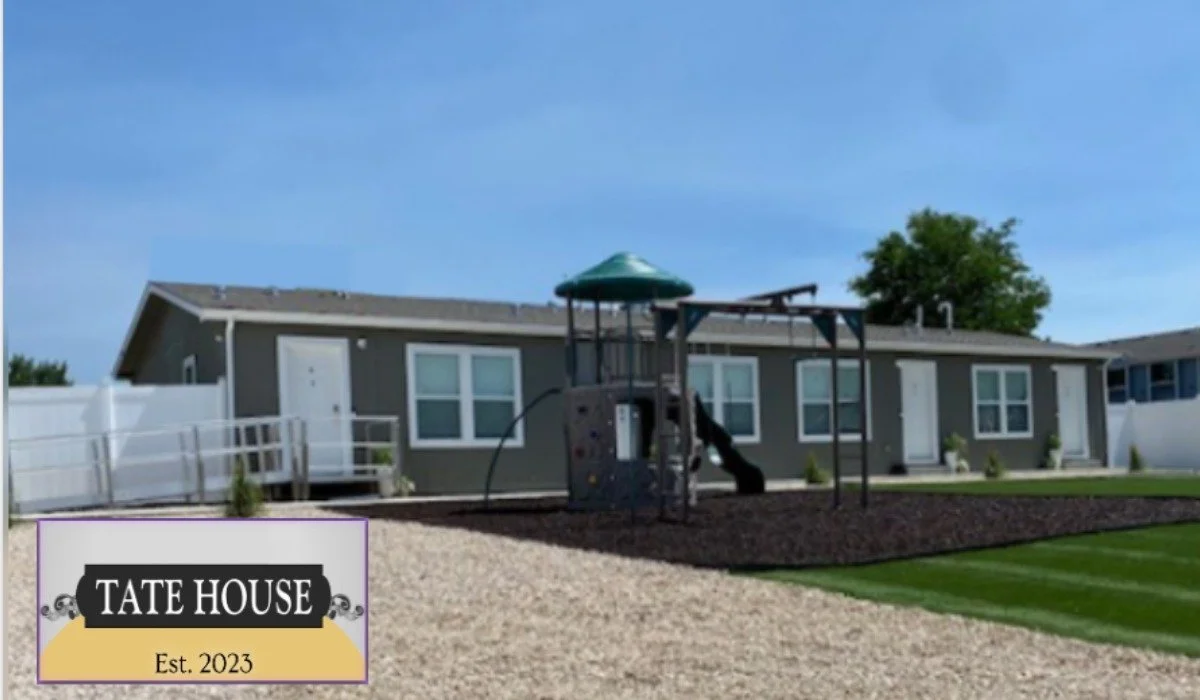 Outside view of the Tate House, a grey single story multi-resident home with a playground in front