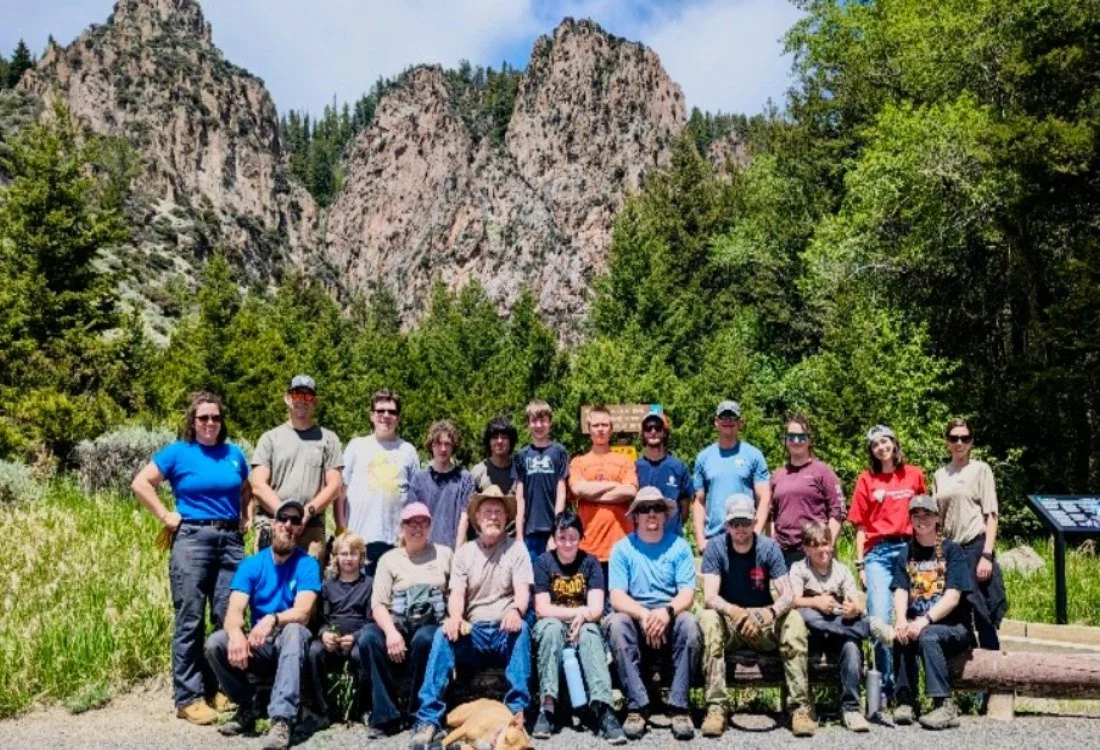 Group photo of youth and adults outside at the Five Springs Falls Campground, posed in front of a tall mountainscape and green trees