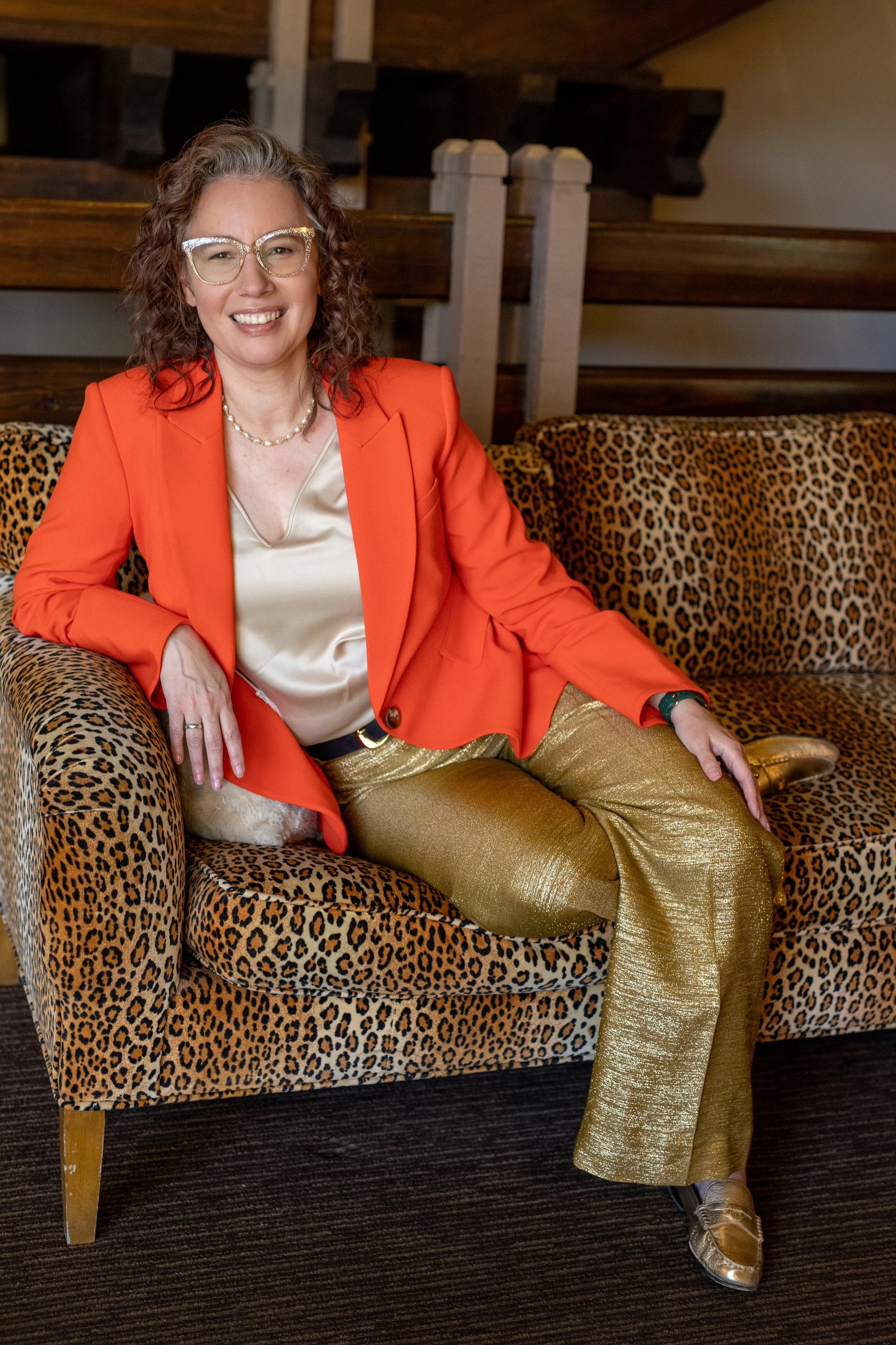 Angela Marie D'Antonio, executive coach for neurodivergent founders and keynote speaker, smiling in an orange blazer and gold pants seated on a leopard-print sofa.