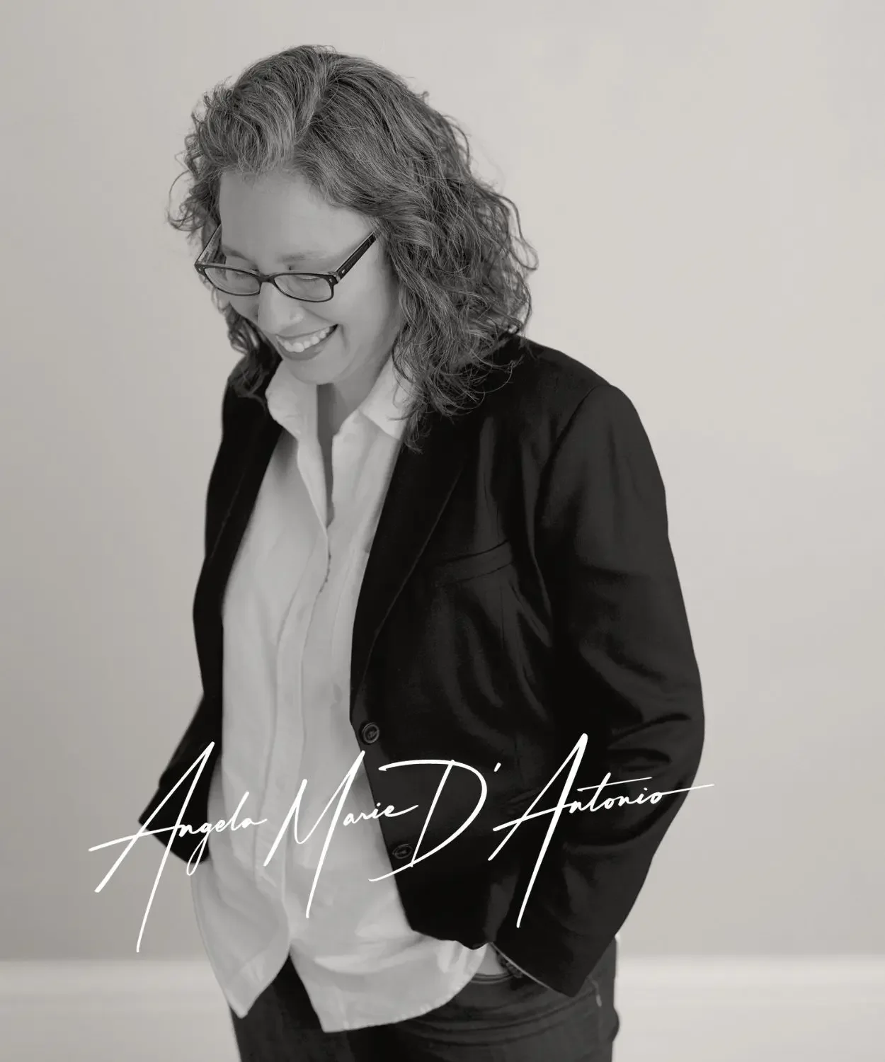 Black-and-white portrait of Angela Marie D’Antonio, education keynote speaker, wearing glasses and a blazer, smiling while looking down.