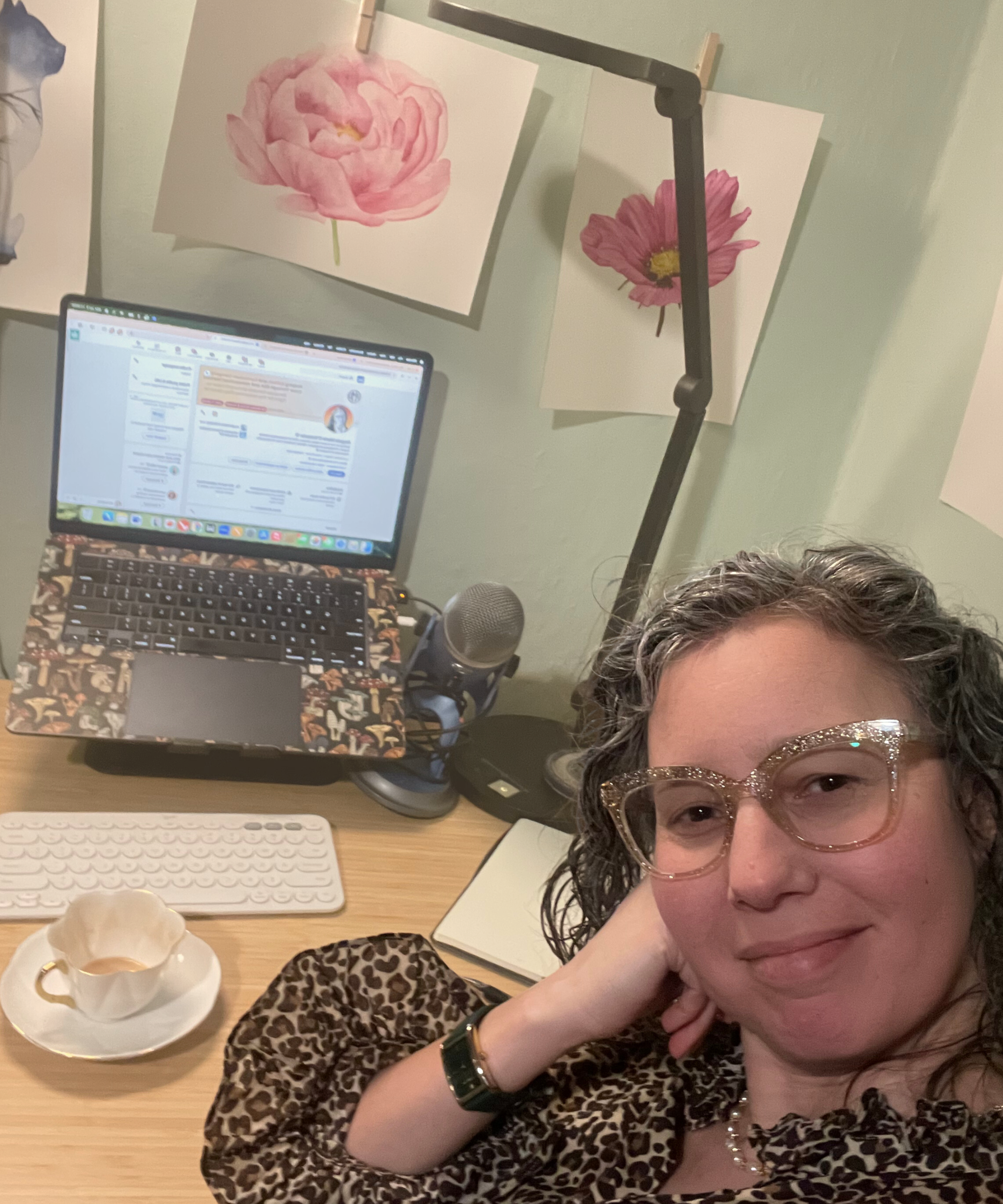 Angela Marie D'Antonio, a woman with curly hair and glasses smiling at the camera, sitting at a desk with a cup of tea, a keyboard, a notebook, and a computer monitor, with floral paintings hanging on the wall behind her.
