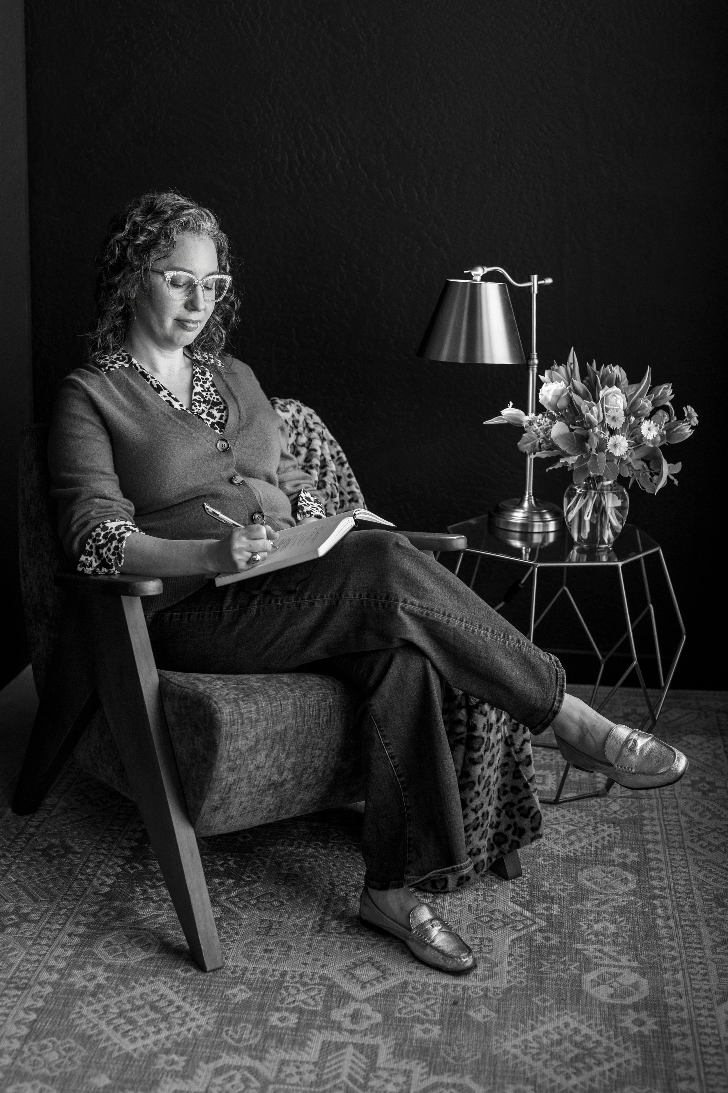 Angela Marie D'Antonio, executive coach for neurodivergent founders and writer on Calm Ambition, seated in a wooden chair writing in a notebook beside a lamp and vase of flowers.