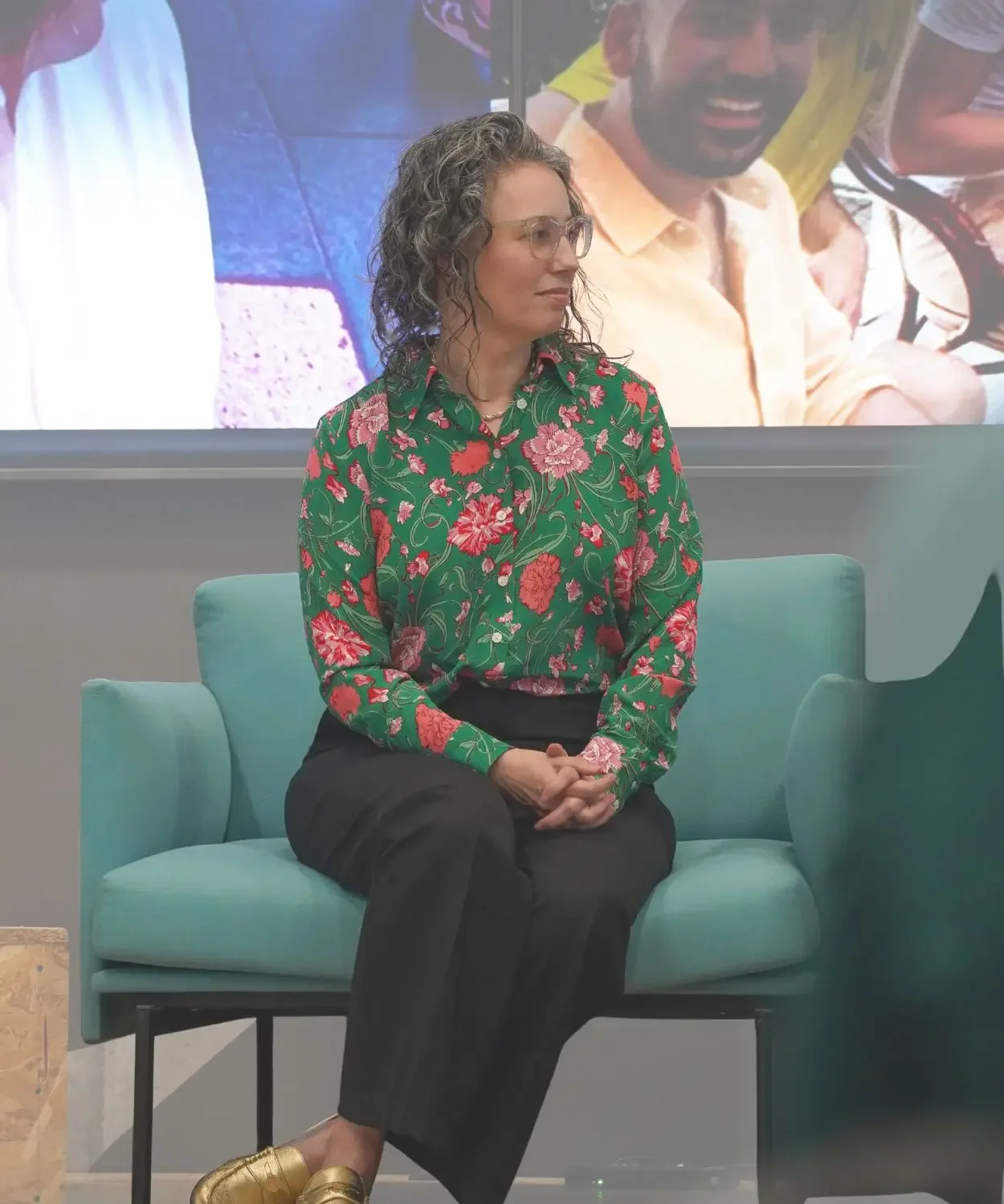 Angela Marie D’Antonio, education keynote speaker, seated on a stage chair, wearing a green floral blouse and black pants, listening attentively during a panel discussion.
