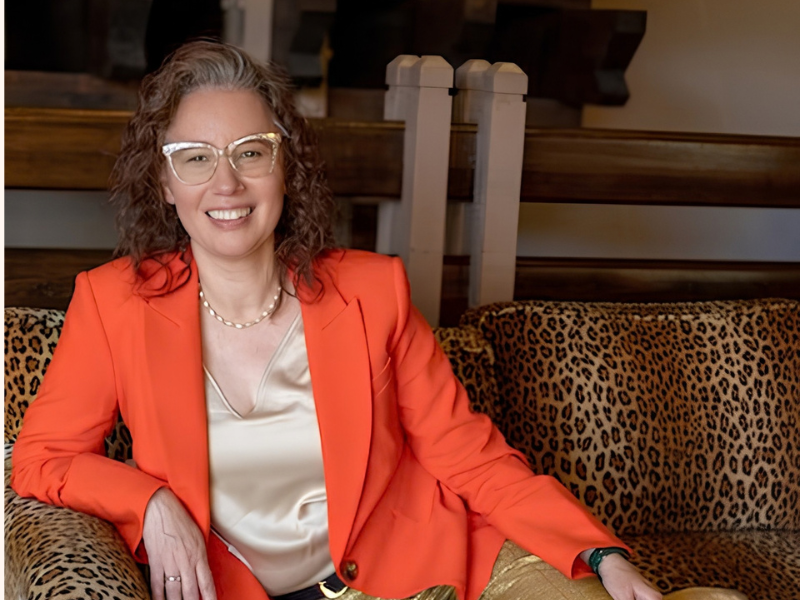 Angela Marie D'Antonio, neurodiversity speaker and advisor, smiles at camera. She is sitting on leopard print couch and has orange and gold clothing including her signature gold glitter glasses.