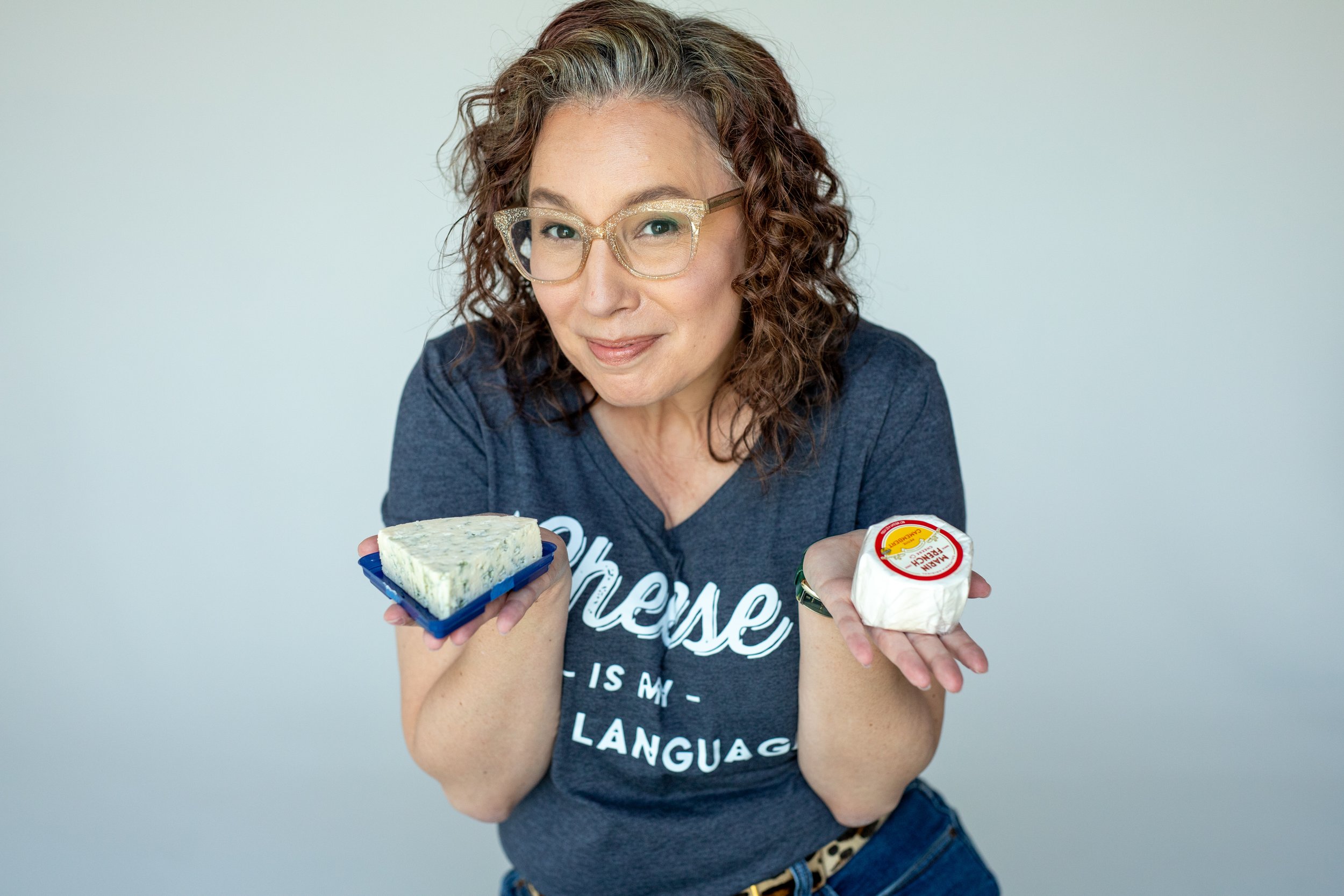 Angela Marie D'Antonio, executive coach for neurodivergent founders and Associate of Cheese, smiling in a "Cheese Is My Love Language" tee while holding a wedge of blue cheese and round of brie.